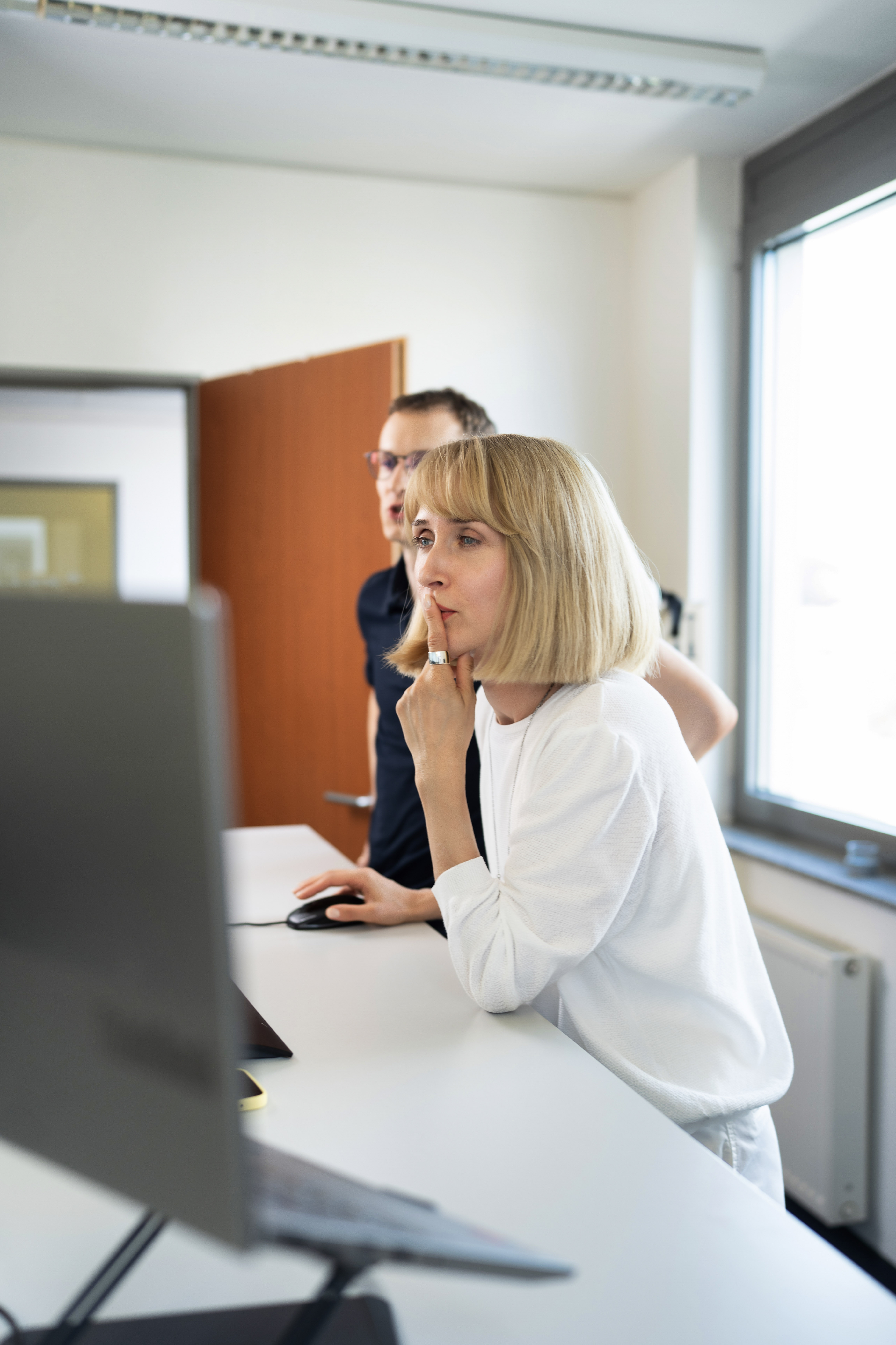 Eine Frau mit blonden, schulterlangen Haaren und einem weißen Oberteil betrachtet nachdenklich einen Computerbildschirm, während ein Mann im Hintergrund ebenfalls auf den Bildschirm schaut. Der Raum ist hell und modern eingerichtet.