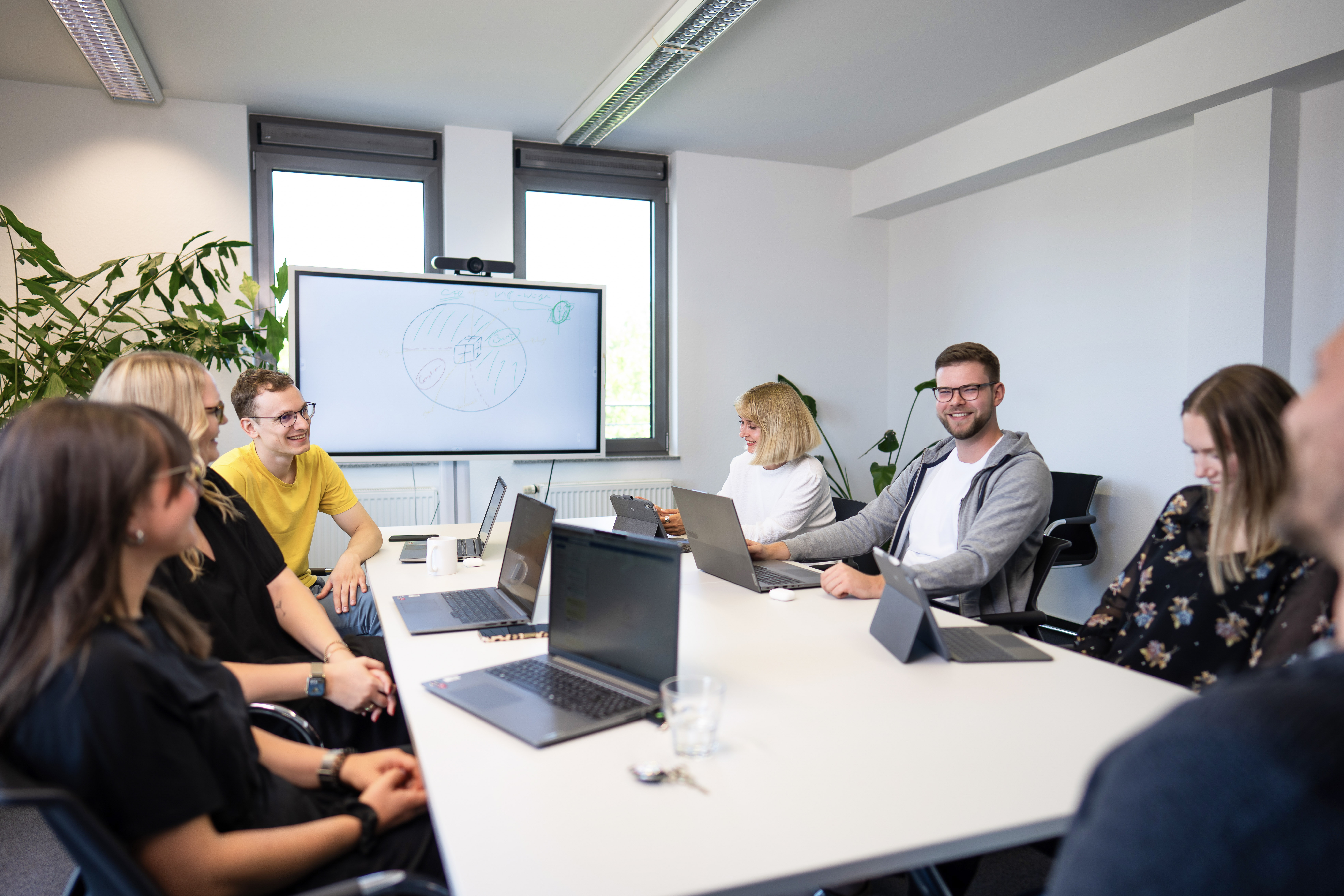 Eine Gruppe von sechs Personen sitzt an einem Tisch in einem modernen Besprechungsraum, während sie an Laptops arbeiten. An der Wand steht ein Whiteboard mit Zeichnungen, und eine Pflanze ist im Hintergrund sichtbar. Alle Teilnehmer wirken engagiert und kommunizieren miteinander.