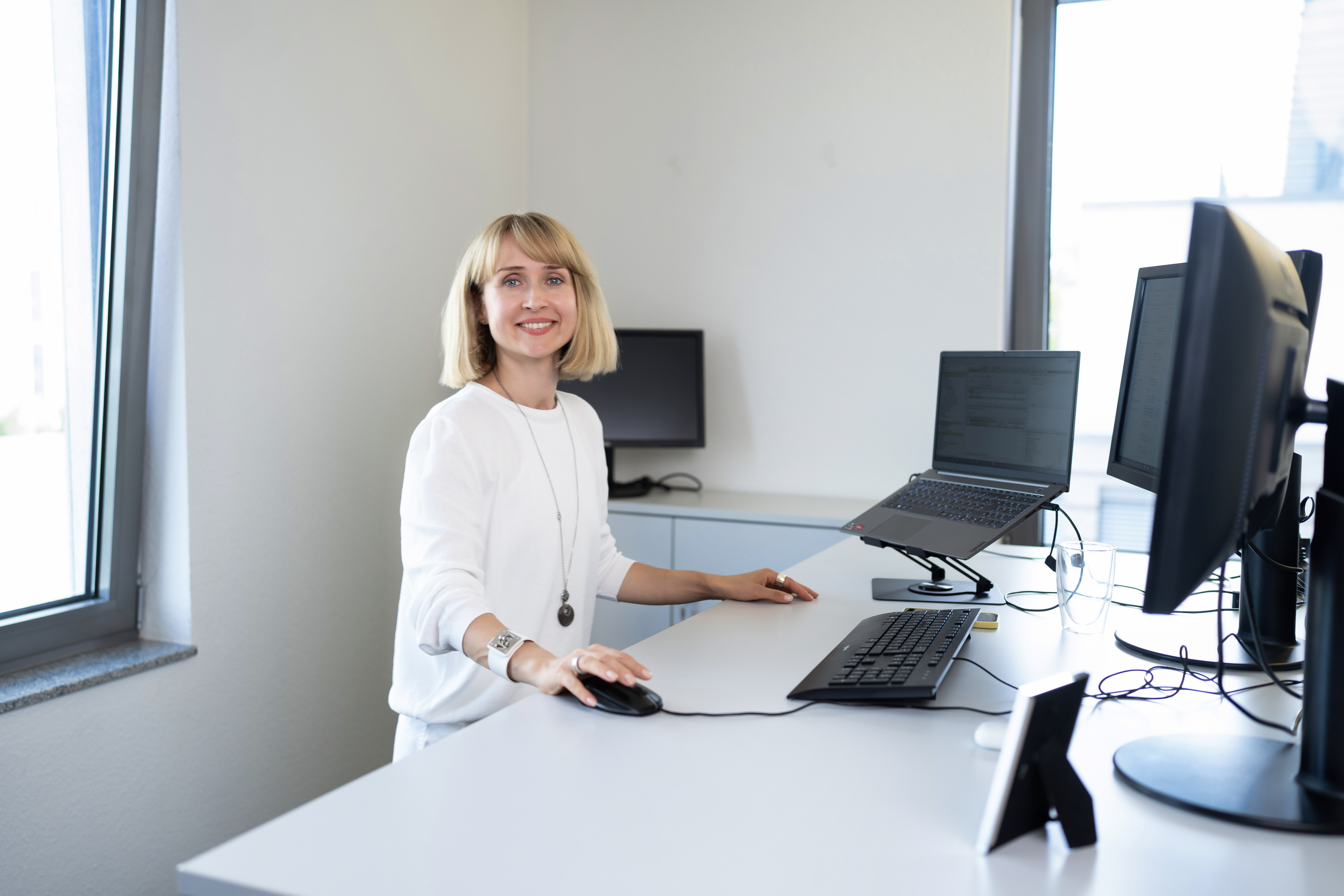 Eine junge Frau mit blonden Haaren steht an einem modernen Schreibtisch und lächelt, während sie mit der Maus an ihrem Computer arbeitet. Auf dem Schreibtisch befinden sich mehrere Monitore und ein Laptop. Die Büroumgebung ist hell und freundlich.