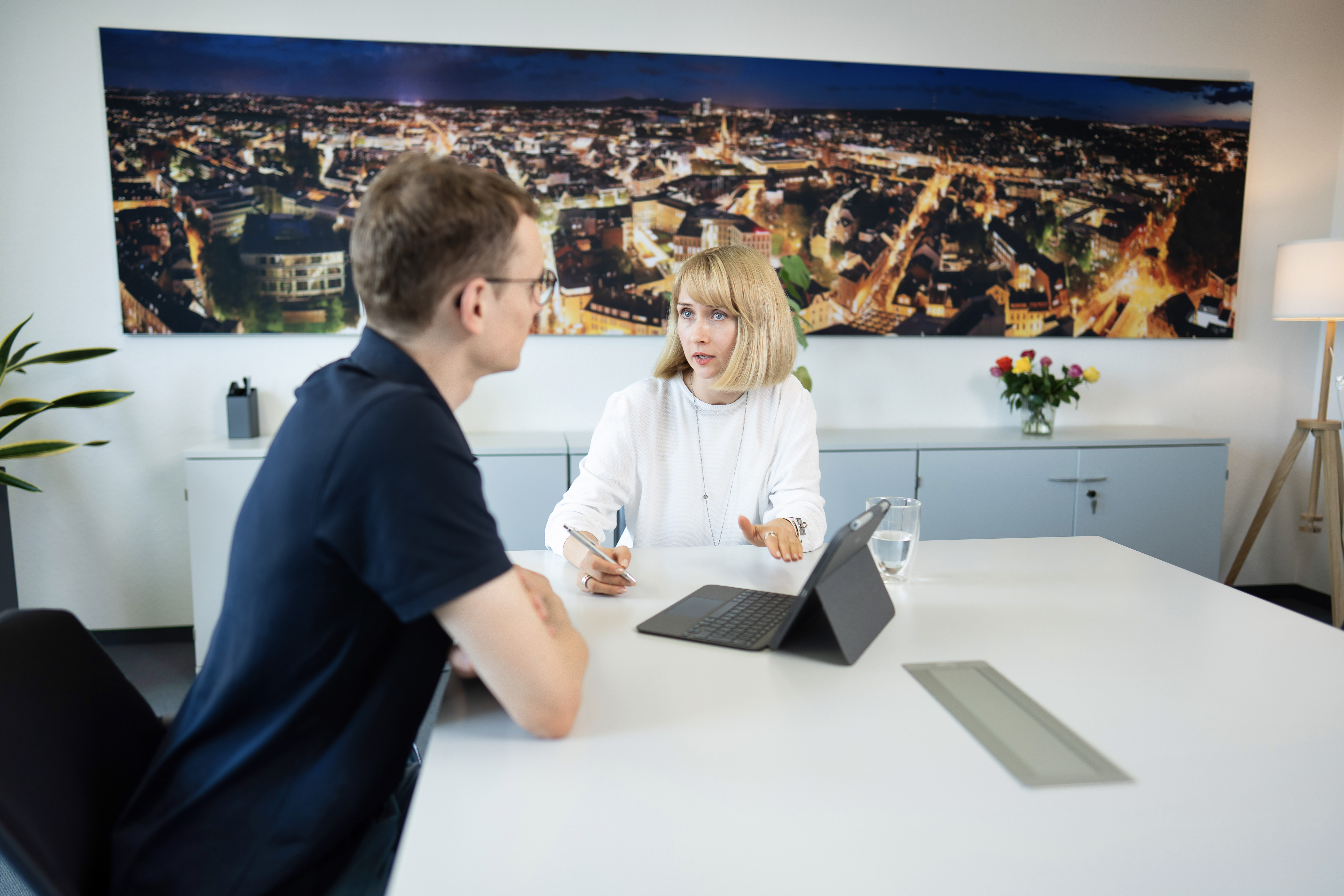 Eine Frau mit blonden Haaren und einem weißen Oberteil sitzt an einem Tisch und spricht mit einem Mann in einem schwarzen T-Shirt; im Hintergrund ist eine städtische Skyline bei Nacht zu sehen. Auf dem Tisch steht ein Glas Wasser und ein Tablet.