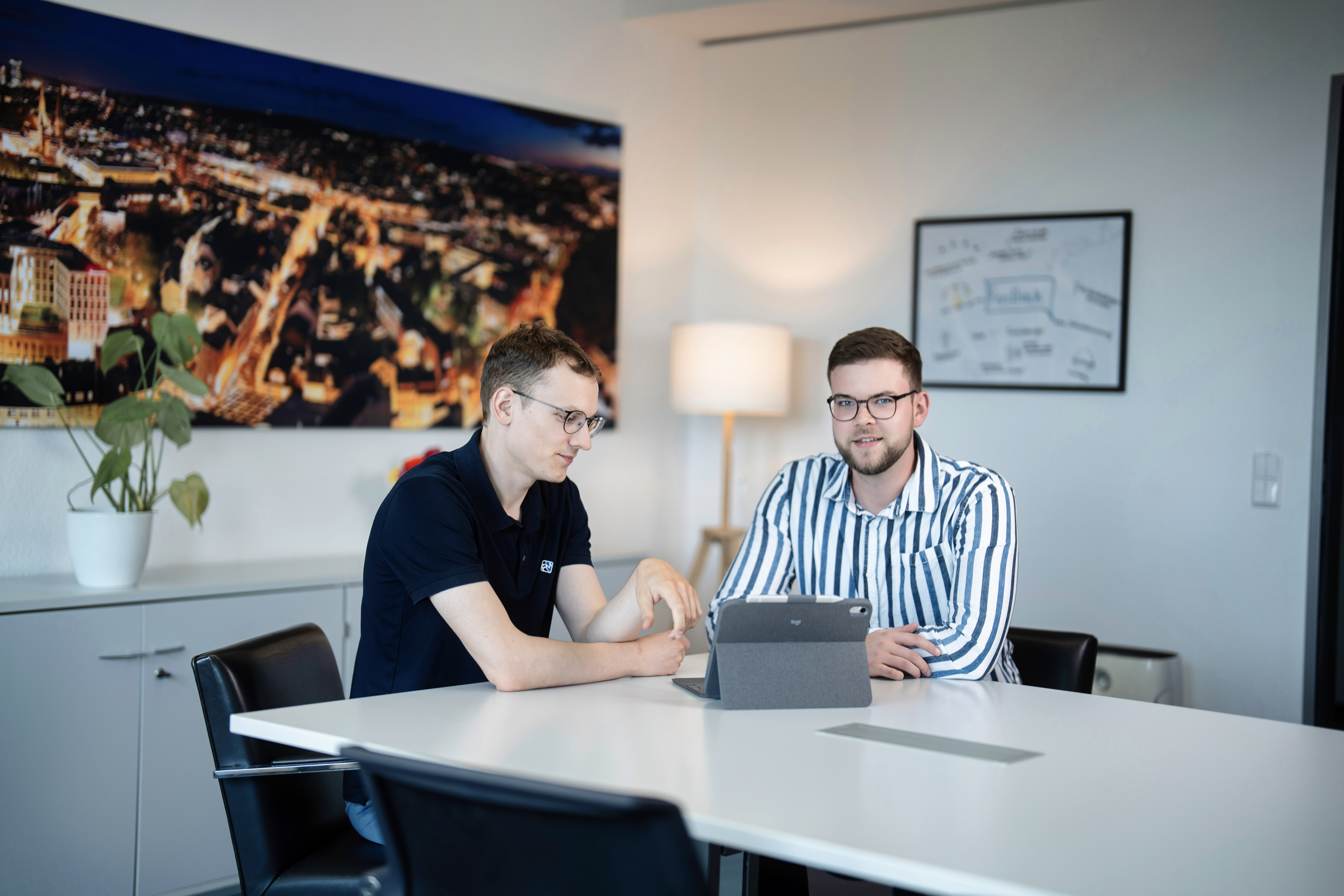 Zwei Männer sitzen an einem Tisch in einem modern eingerichteten Büro, während sie auf ein Tablet schauen. Im Hintergrund ist ein großes Stadtbild und ein Flipchart zu sehen.