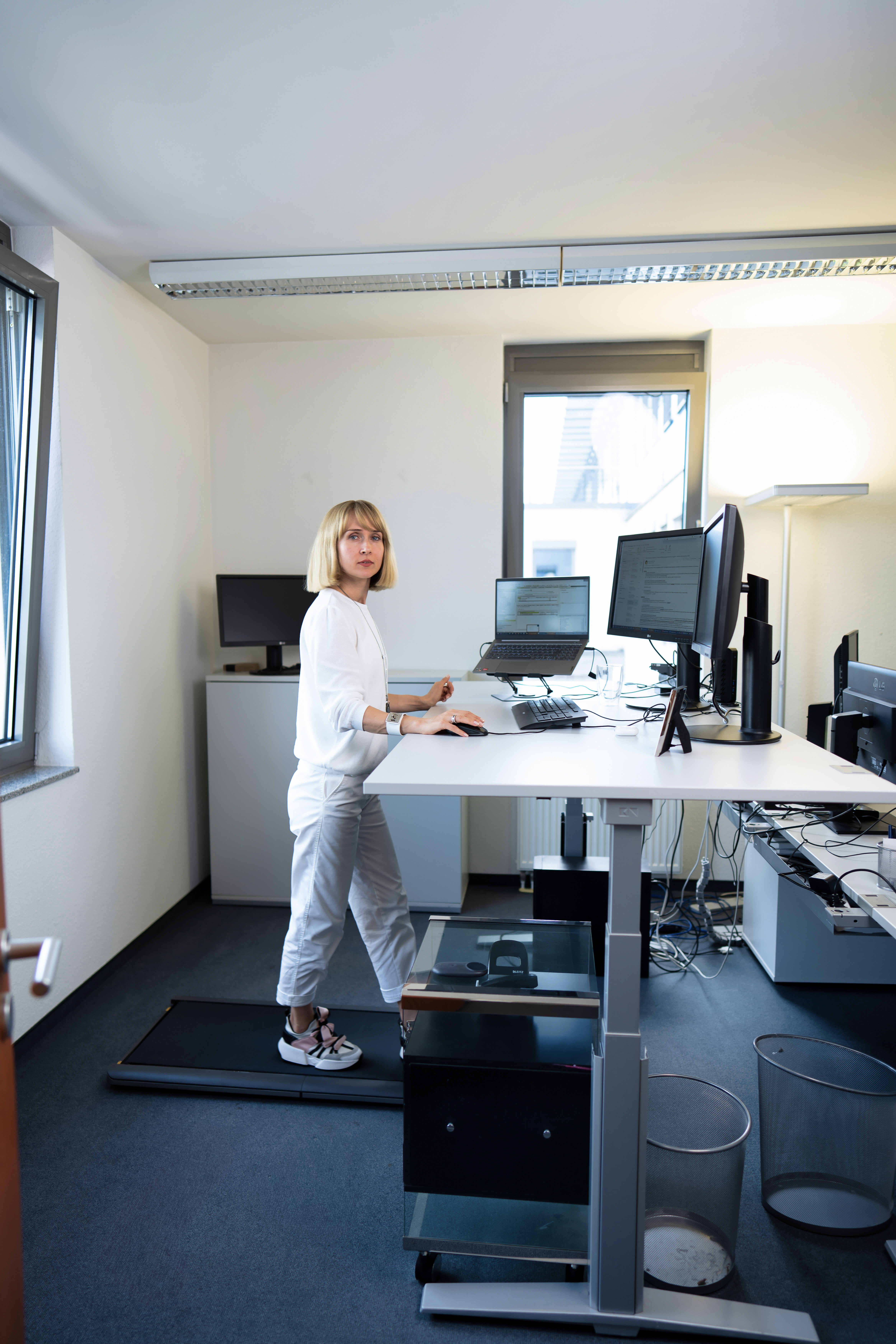 Eine Frau mit blonden, schulterlangen Haaren steht an einem höhenverstellbaren Schreibtisch in einem modernen Büro, während sie an einem Computer arbeitet. Der Raum ist hell und minimalistisch eingerichtet, mit mehreren Bildschirmen und Büroausstattung im Hintergrund.