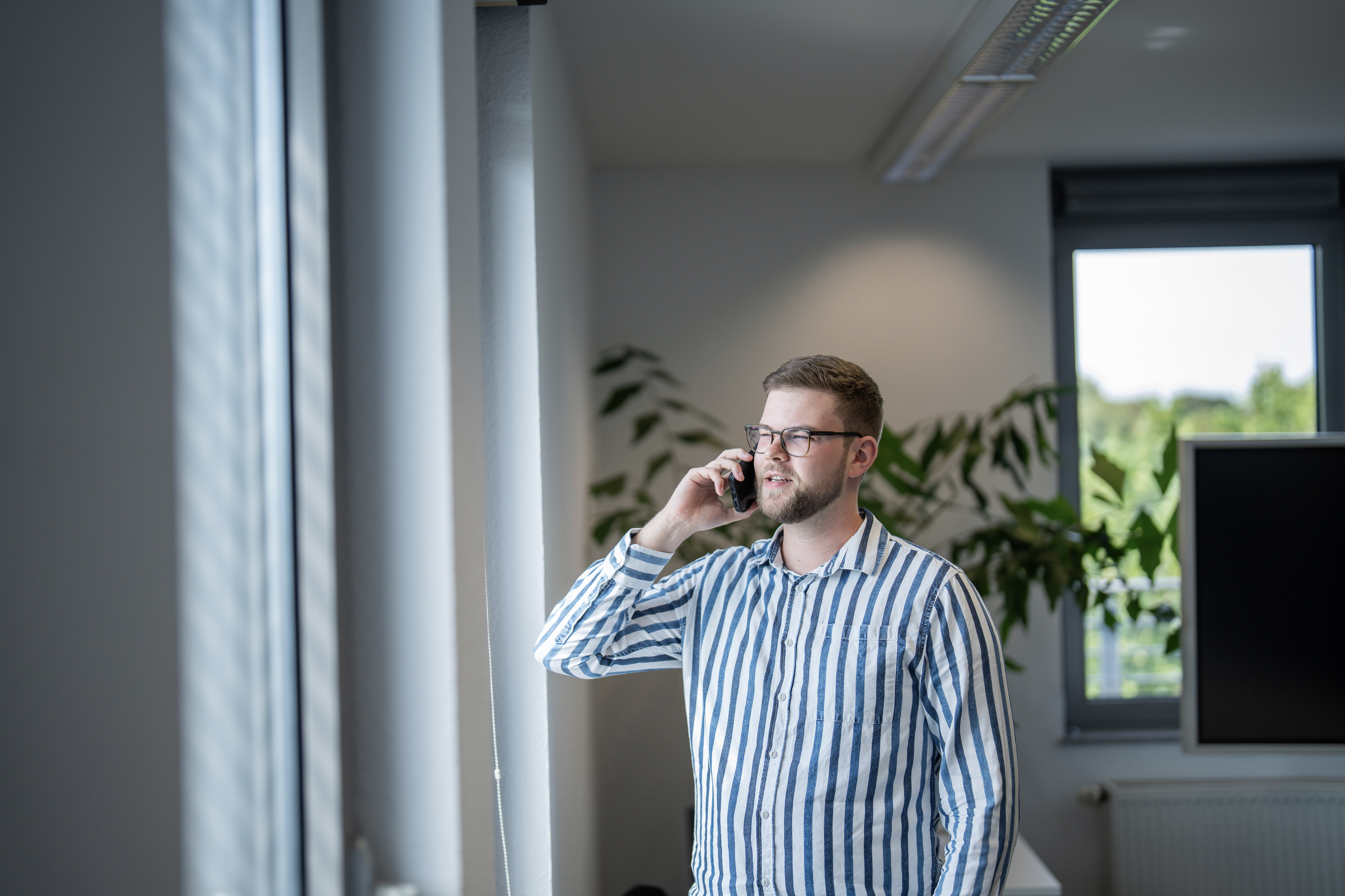 Ein Mann mit Brille trägt ein gestreiftes Hemd und telefoniert in einem modernen Büro. Im Hintergrund sind große Fenster und Pflanzen zu sehen.