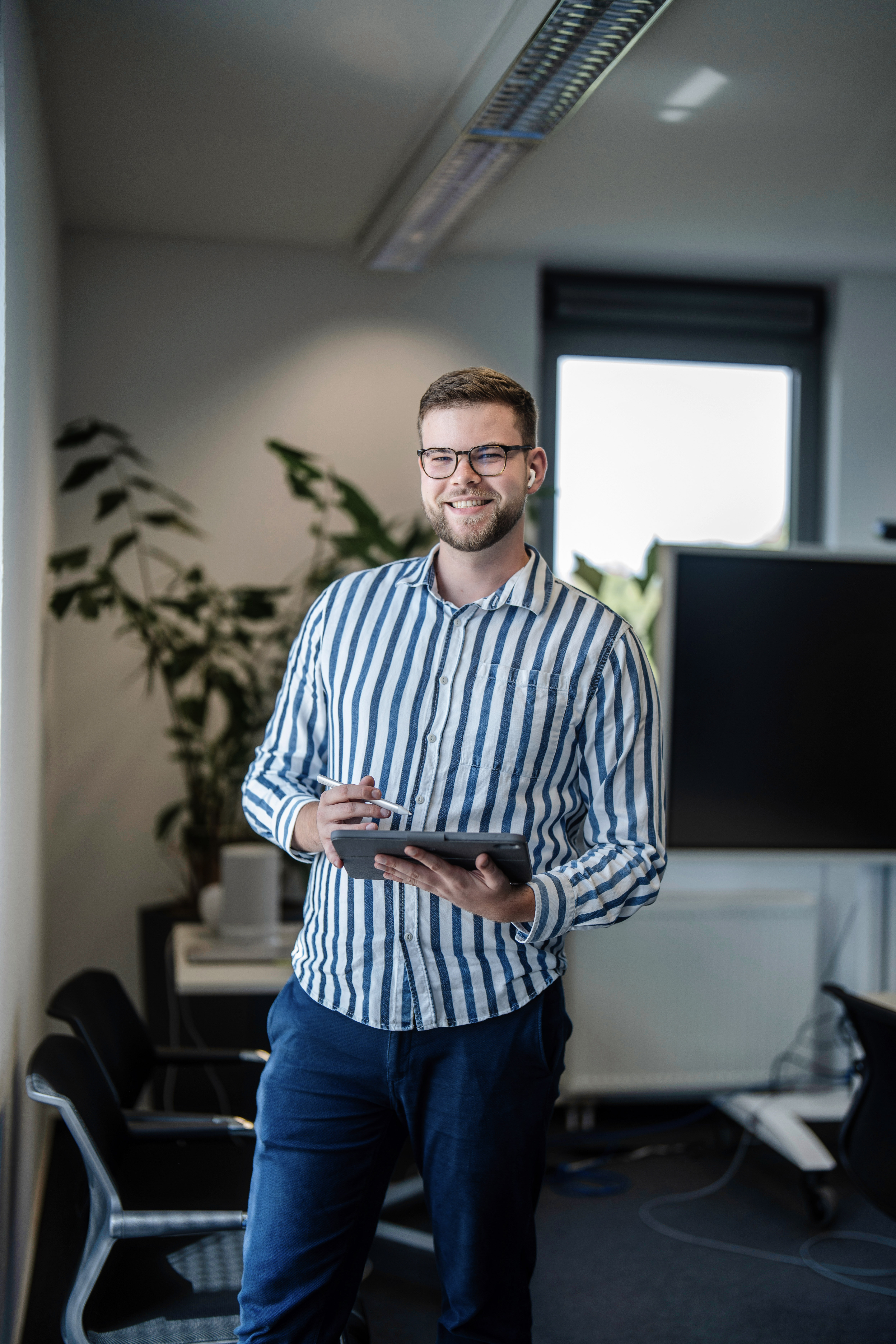 Ein junger Mann mit Brille steht in einem modernen Büro und hält ein Tablet in der Hand, während er in die Kamera lächelt. Im Hintergrund sind Pflanzen und ein Computerbildschirm zu sehen.