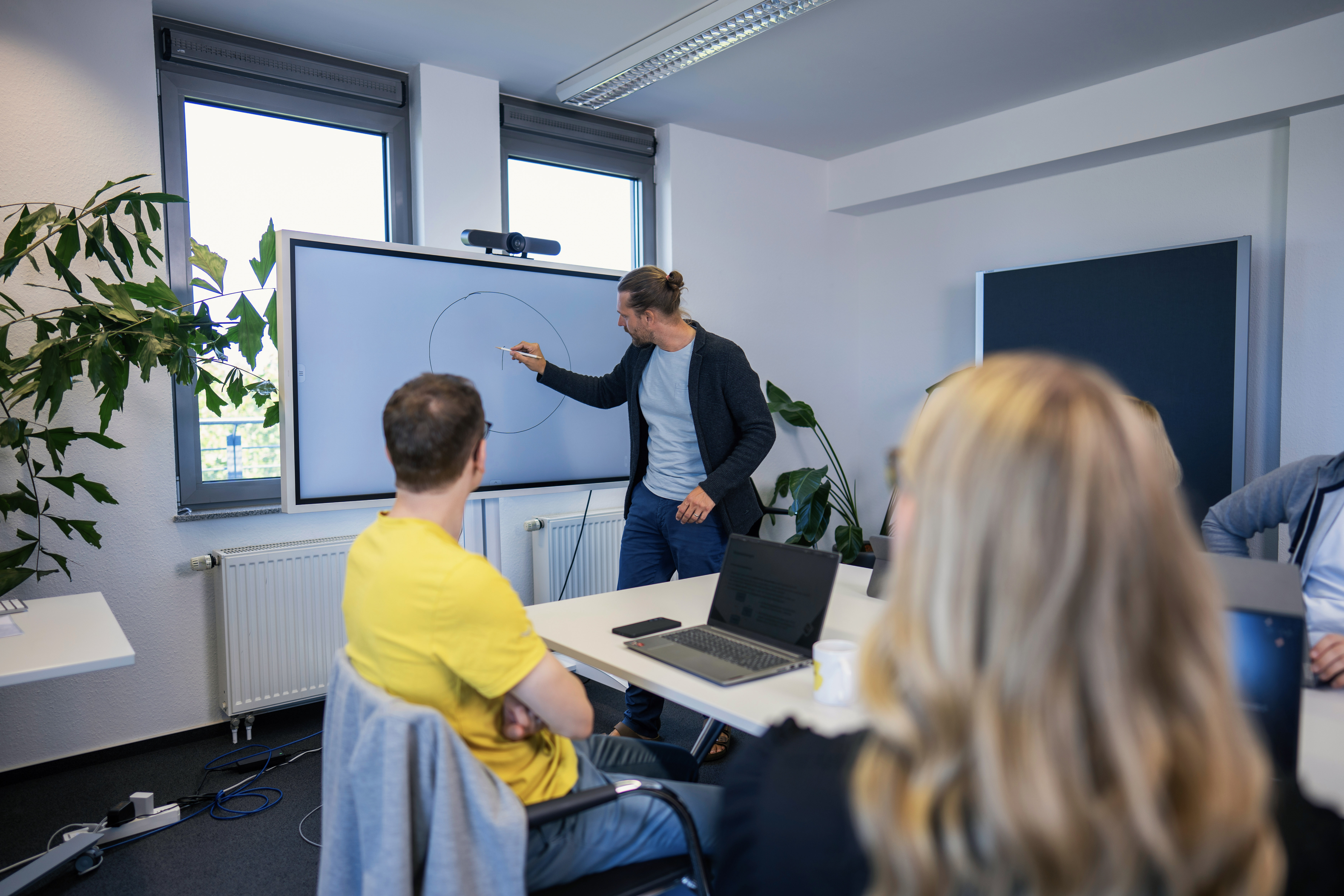 Ein Mann zeichnet einen Kreis auf einem digitalen Bildschirm während einer Präsentation in einem modernen Konferenzraum. Im Vordergrund sitzen mehrere Personen, die ihm aufmerksam zusehen. Pflanzen und Fenster mit Tageslicht sind im Hintergrund sichtbar.