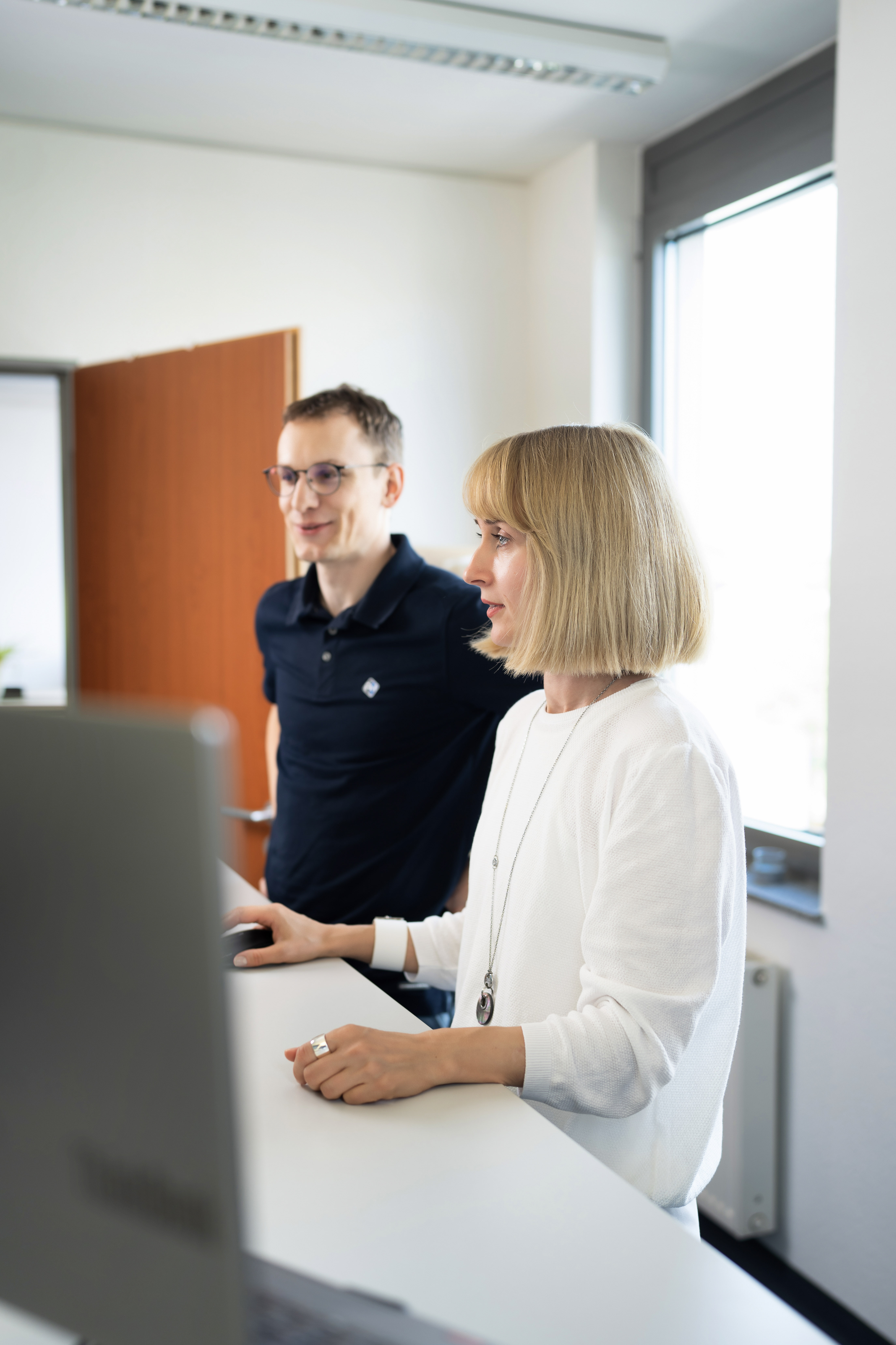 Ein Mann und eine Frau stehen nebeneinander und schauen auf einen Computerbildschirm. Der Mann trägt ein schwarzes Polohemd und hat eine Brille auf, während die Frau ein weißes Oberteil trägt und kurze, blonde Haare hat. Im Hintergrund sind Fenster und eine Holztür zu sehen.