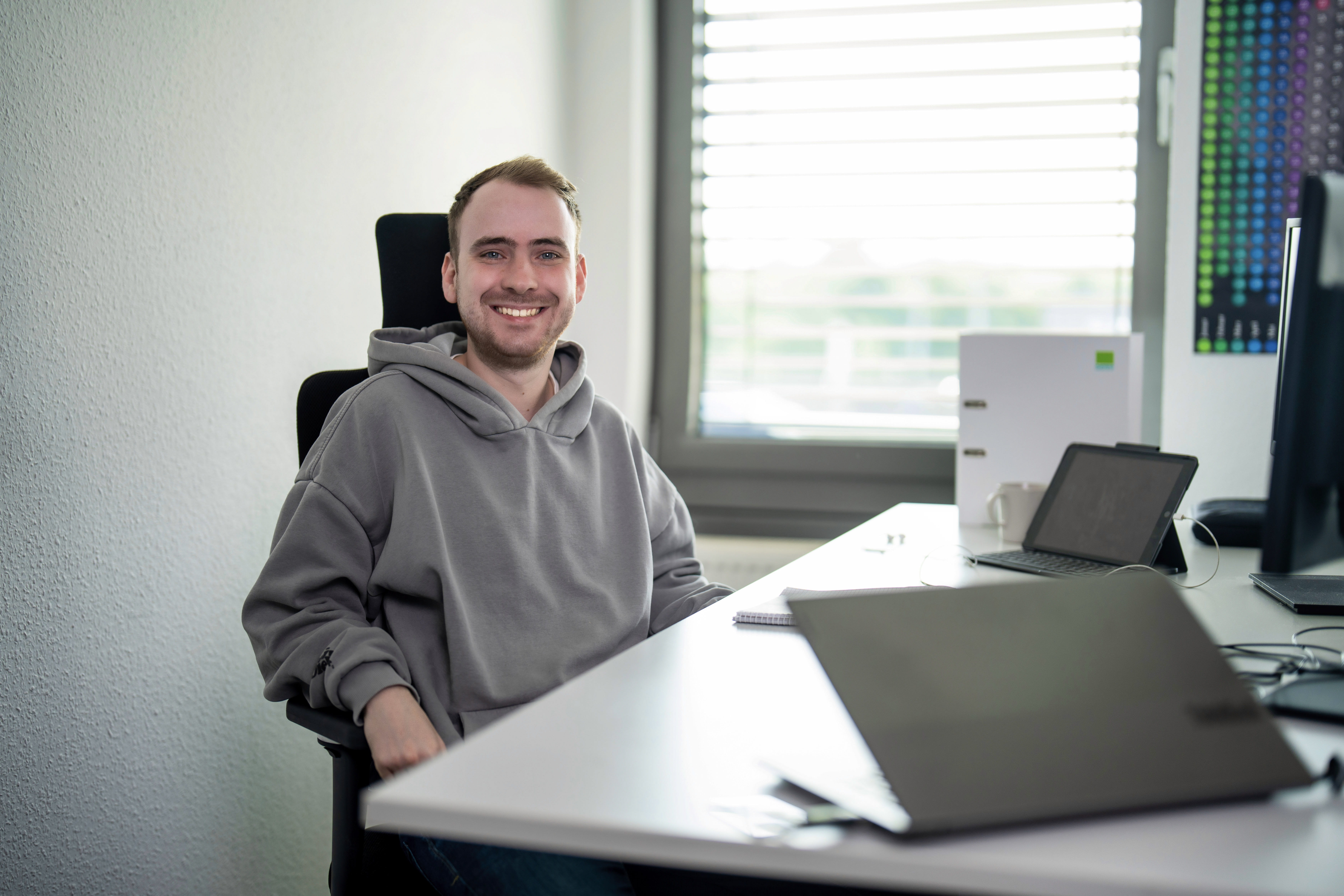 Ein junger Mann mit einem grauen Hoodie sitzt lächelnd an einem Schreibtisch, der mit einem Laptop, einem Notizbuch und einem Monitor ausgestattet ist. Hinter ihm ist ein Fenster mit Jalousien, das Tageslicht in den Raum lässt.