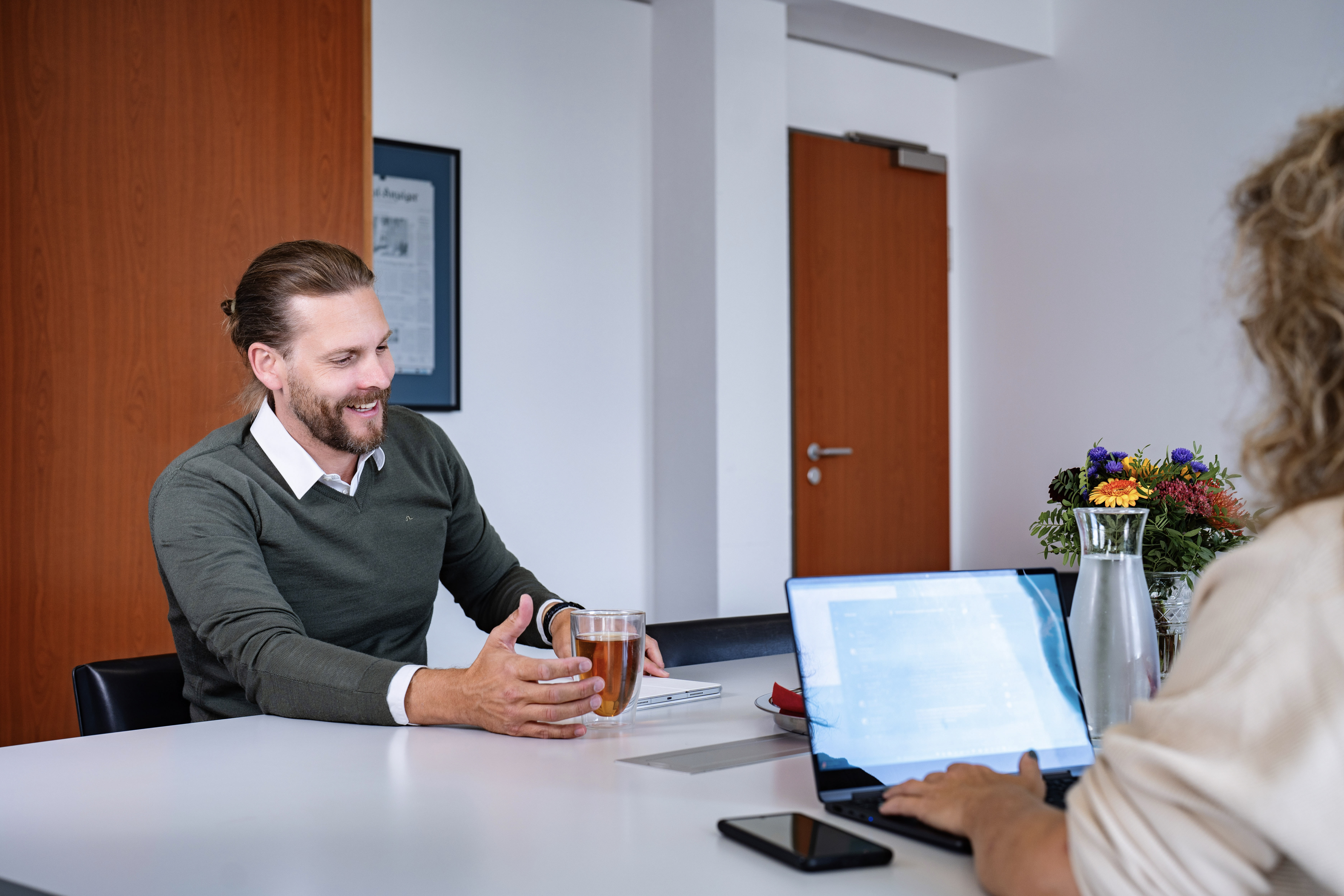 Ein Mann mit leichtem Bart sitzt an einem Tisch und hält ein Glas mit Getränk in der Hand, während er mit einer Frau spricht, die an einem Laptop arbeitet. Im Hintergrund sind Blumen und eine Wasserkaraffe zu sehen.