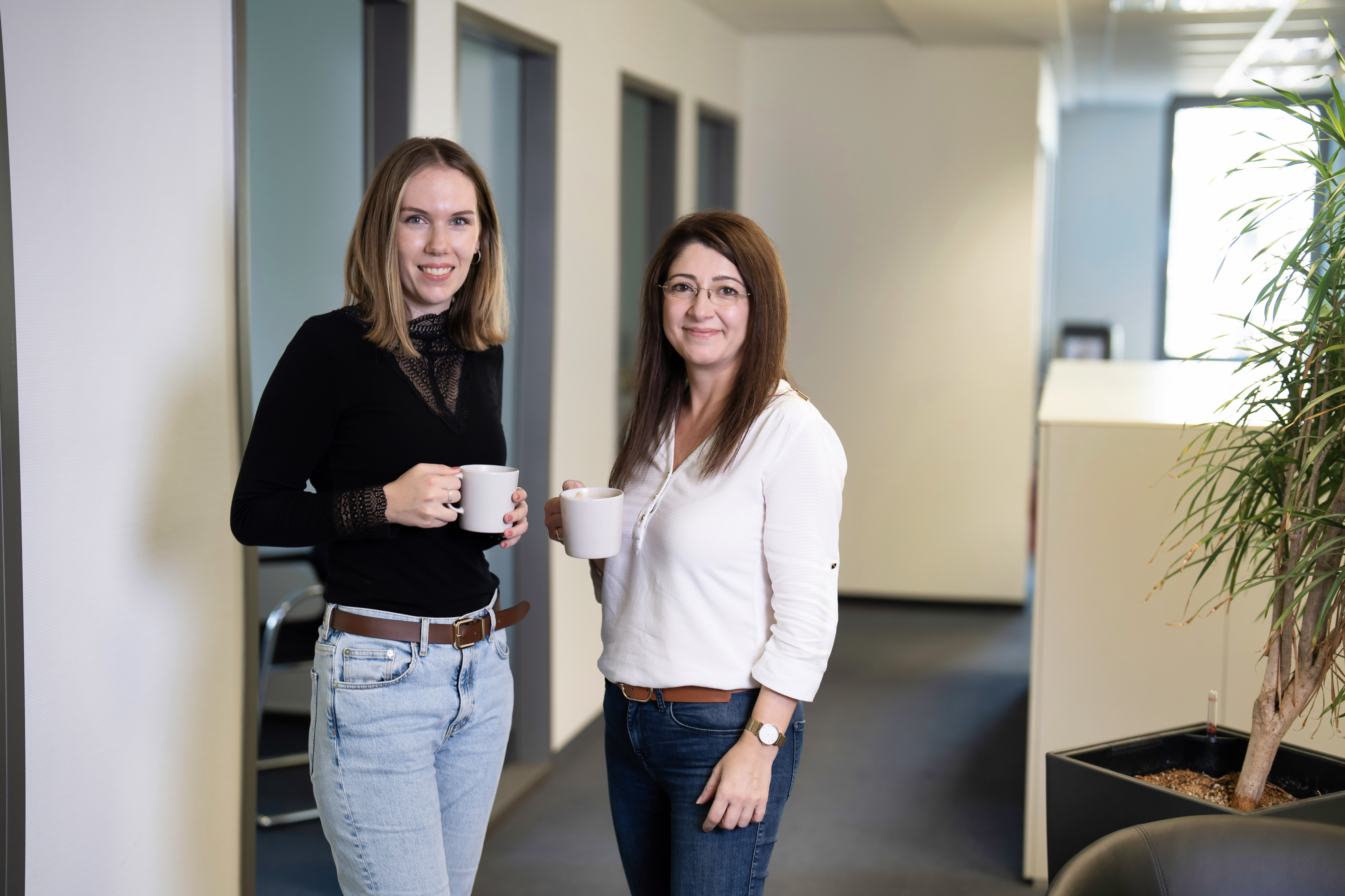 Zwei Frauen stehen in einem modernen Büroumfeld auf einem Flur und halten jeweils eine Tasse in der Hand. Die Frau links trägt ein schwarzes Oberteil und Jeans, während die Frau rechts ein weißes Oberteil und eine braune Hose trägt. Im Hintergrund sind Bürotüren und ein Pflanzenkübel sichtbar.