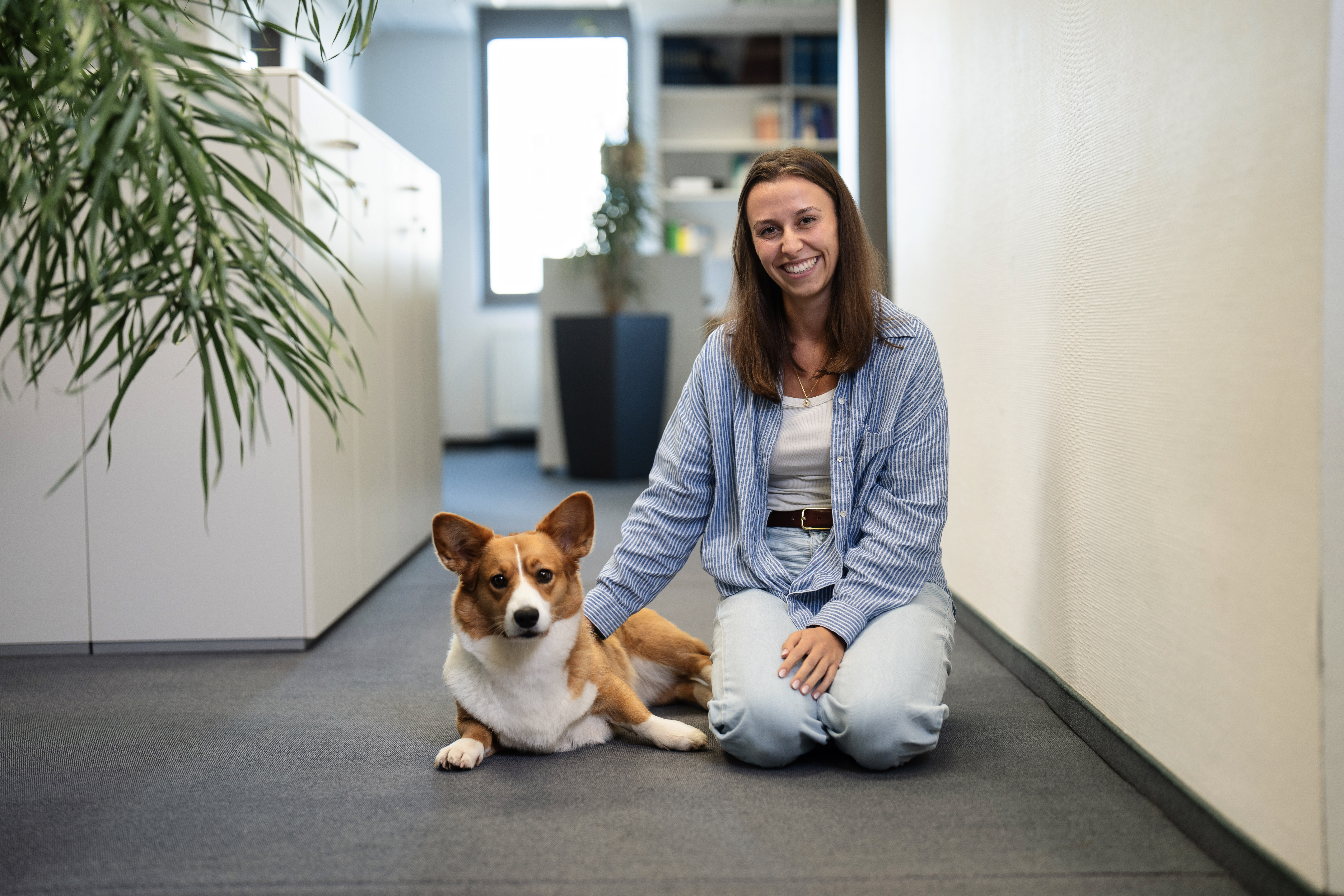Eine Frau sitzt im Büro auf dem Boden und streichelt einen Hund, der neben ihr liegt. Im Hintergrund sind Regale und Pflanzen sichtbar.