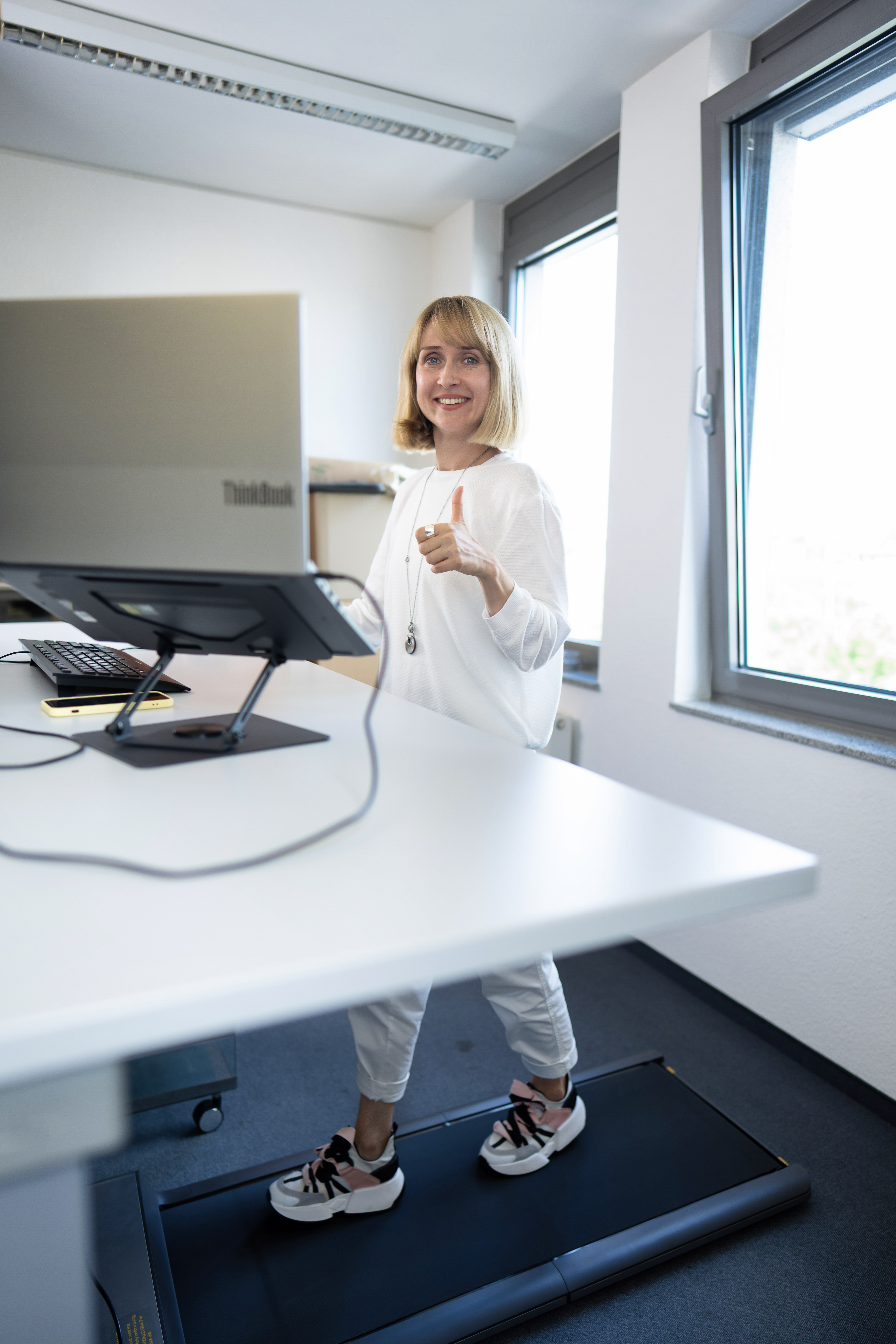Eine Frau steht lächelnd auf einem Laufband in einem hellen Büro, während sie an einem Computer arbeitet und Daumen nach oben zeigt.