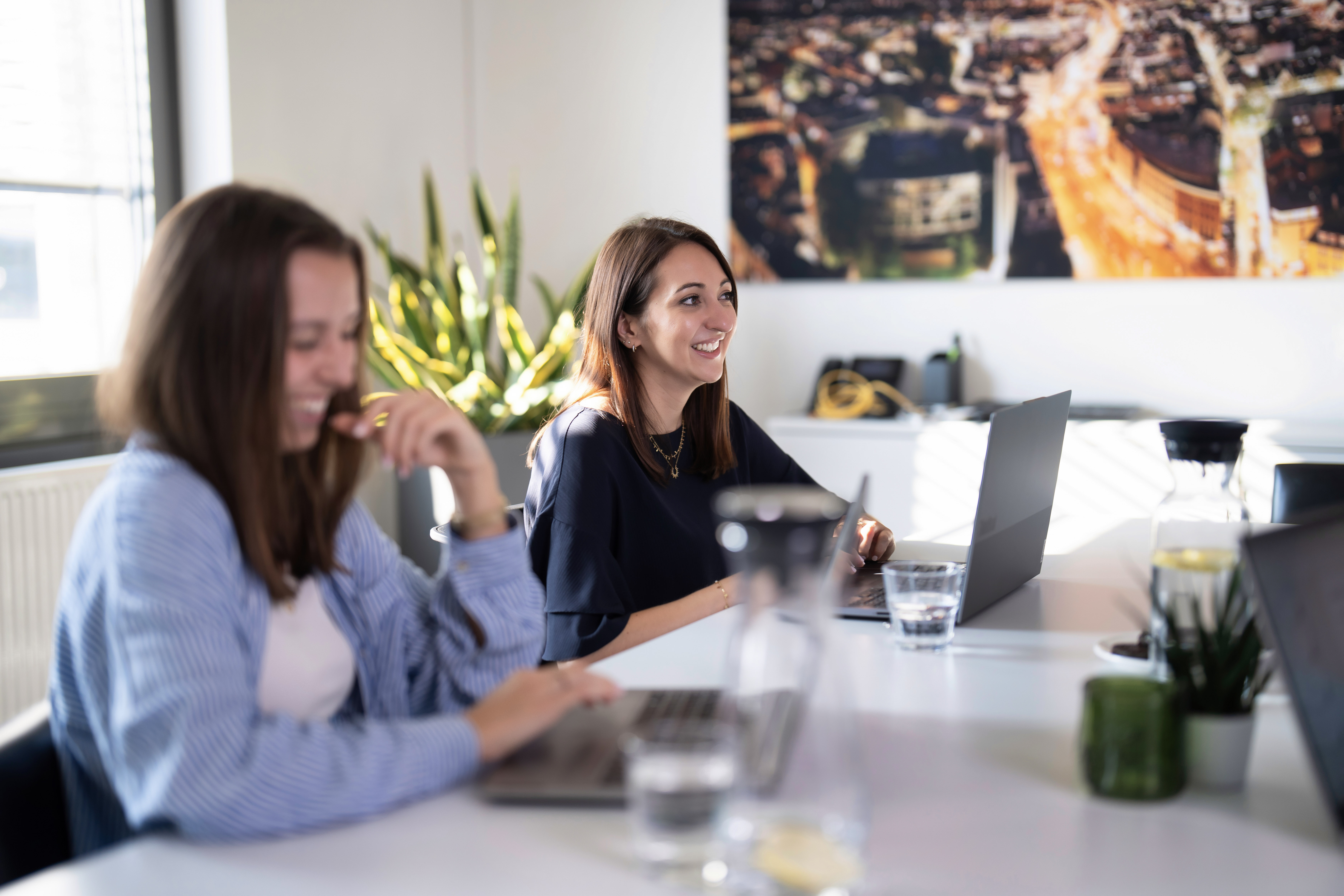 Zwei Frauen sitzen an einem Tisch in einem modern eingerichteten Büroraum; eine Frau lächelt, während die andere lachend auf ihrem Laptop arbeitet. Im Hintergrund ist ein großes Bild mit einer Stadtansicht zu sehen. Auf dem Tisch stehen Wassergläser und Pflanzen.