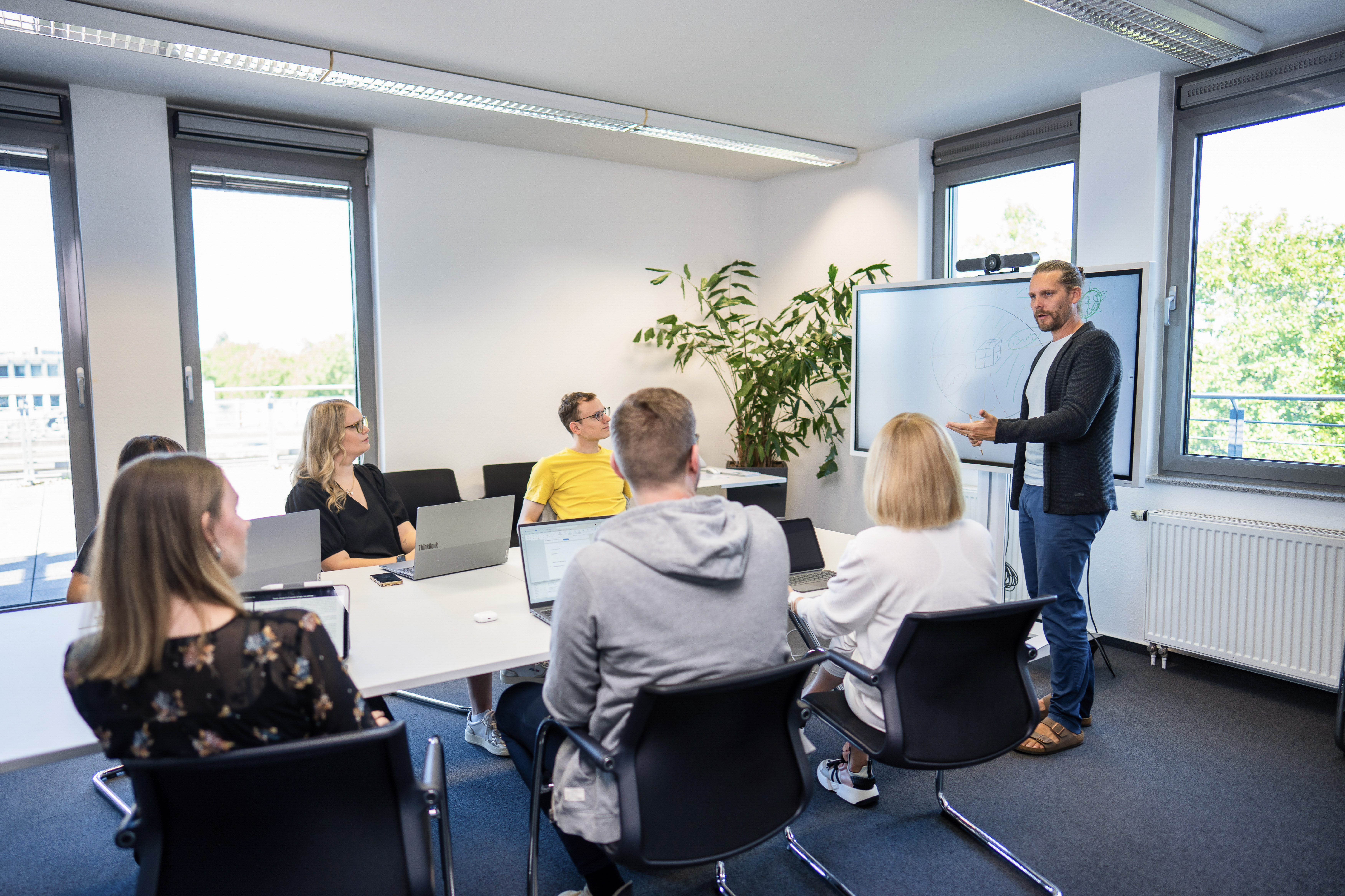 Eine Gruppe von sieben Personen sitzt an einem Konferenztisch in einem hellen Meetingraum. Der Vortragende, ein Mann, steht vor einem Whiteboard und präsentiert während einer Besprechung. Alle Teilnehmer nutzen Laptops und sind aufmerksam. Im Hintergrund ist eine große Pflanze und Fenster mit Tageslicht zu sehen.