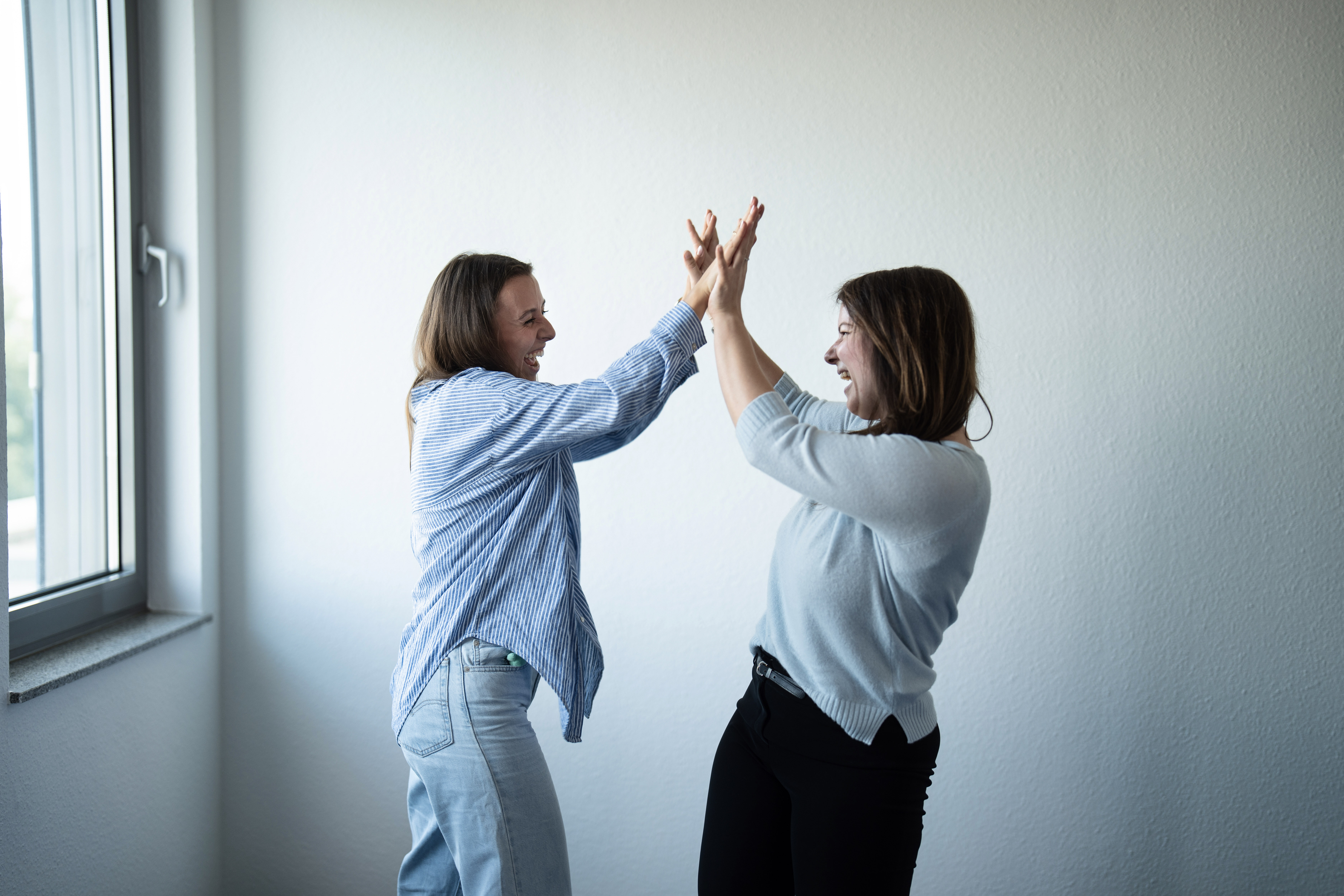 Zwei Frauen stehen sich gegenüber und klatschen einander in die Hände, während sie lachen und offensichtlich Spaß haben. Der Hintergrund ist eine helle Wand und ein Fenster, das natürliches Licht hereinlässt.