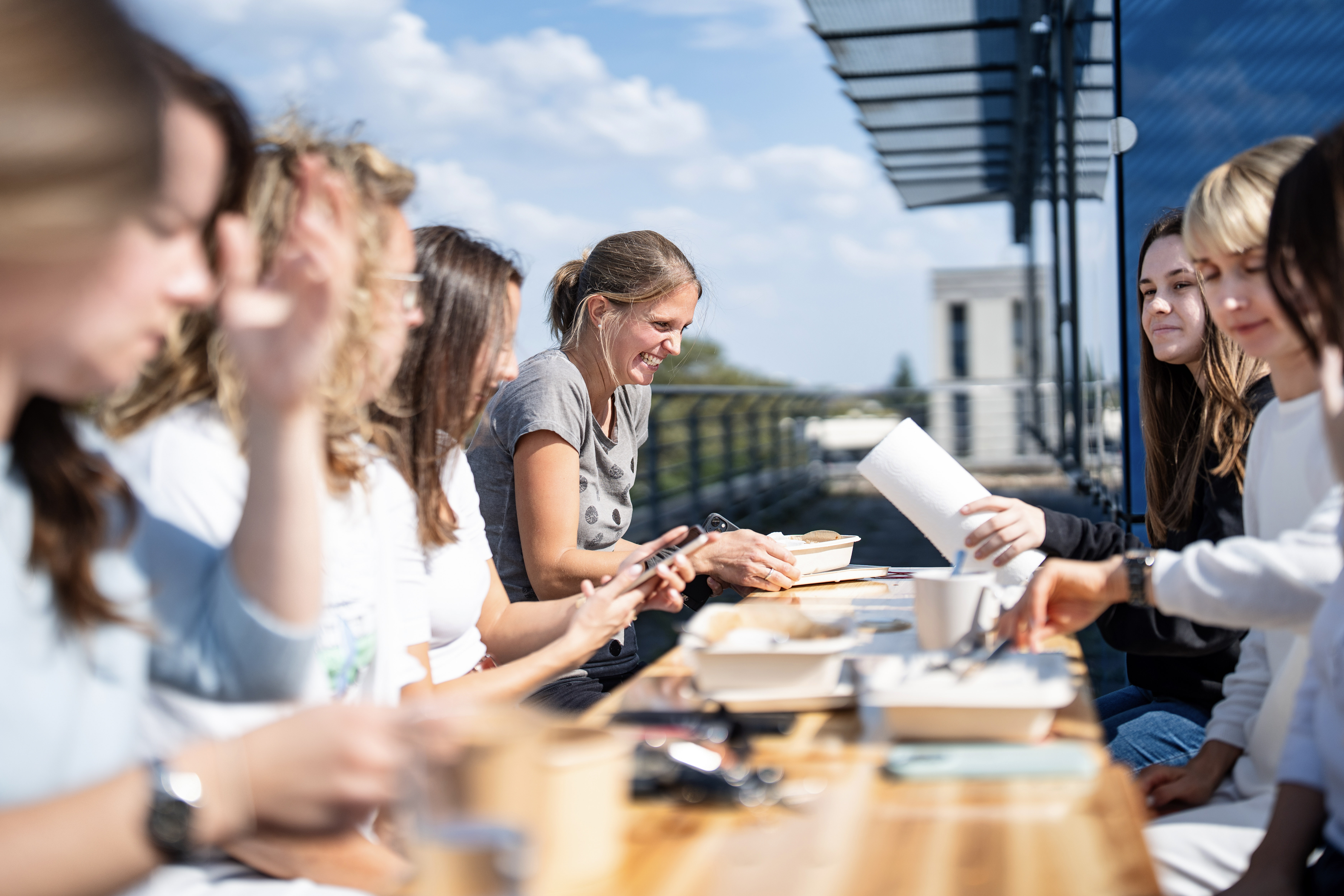 Eine Gruppe von Frauen sitzt an einem langen Tisch im Freien und interagiert freundlich miteinander, während sie an verschiedenen Projekten oder Aktivitäten arbeiten. Die Sonne scheint, und im Hintergrund sind moderne Gebäude und ein blauer Himmel zu sehen.