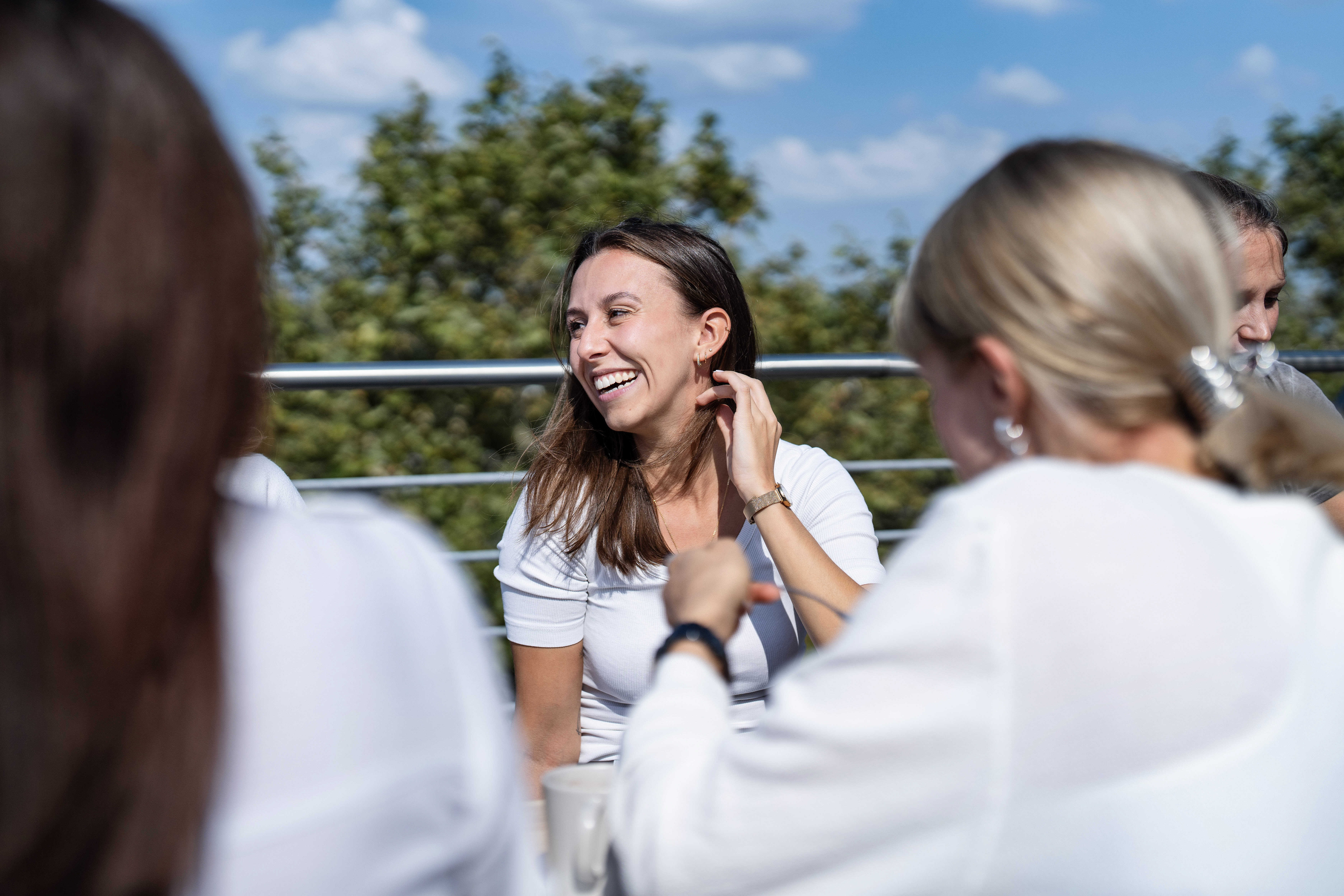 Eine lächelnde Frau mit langen braunen Haaren sitzt an einem Tisch im Freien, umgeben von Personen, die teilweise im Bild sichtbar sind, während sie in einem entspannten Gespräch vertieft sind. Im Hintergrund ist ein blauer Himmel mit einigen Wolken und grüne Bäume zu sehen.
