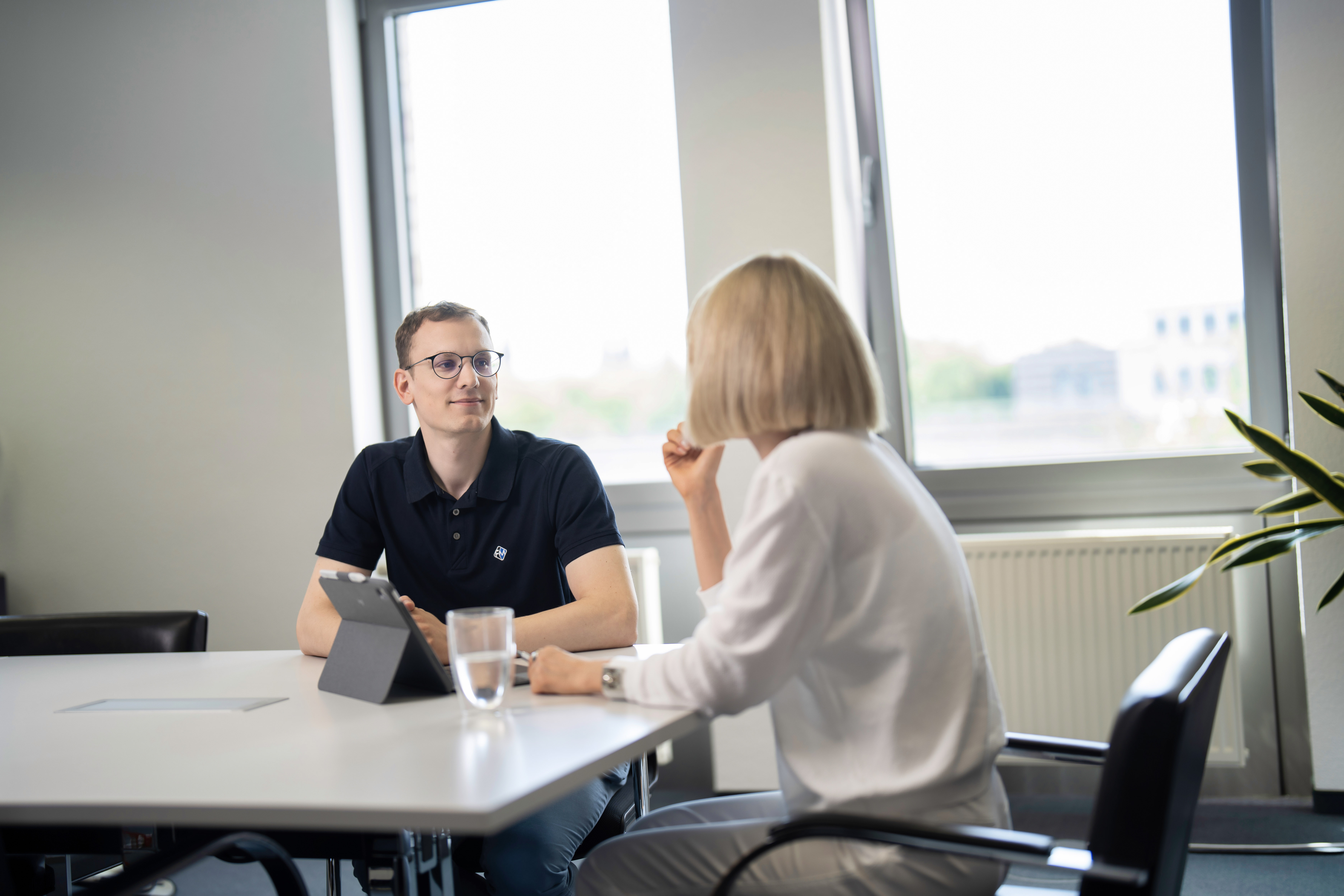 Ein Mann in einem schwarzen Poloshirt sitzt an einem Tisch und schaut eine Frau an, die ihm gegenüber sitzt. Die Frau trägt ein weißes Oberteil und spricht. Auf dem Tisch steht ein Glas Wasser und ein Tablet. Im Hintergrund sind Fenster mit Blick auf eine städtische Landschaft.