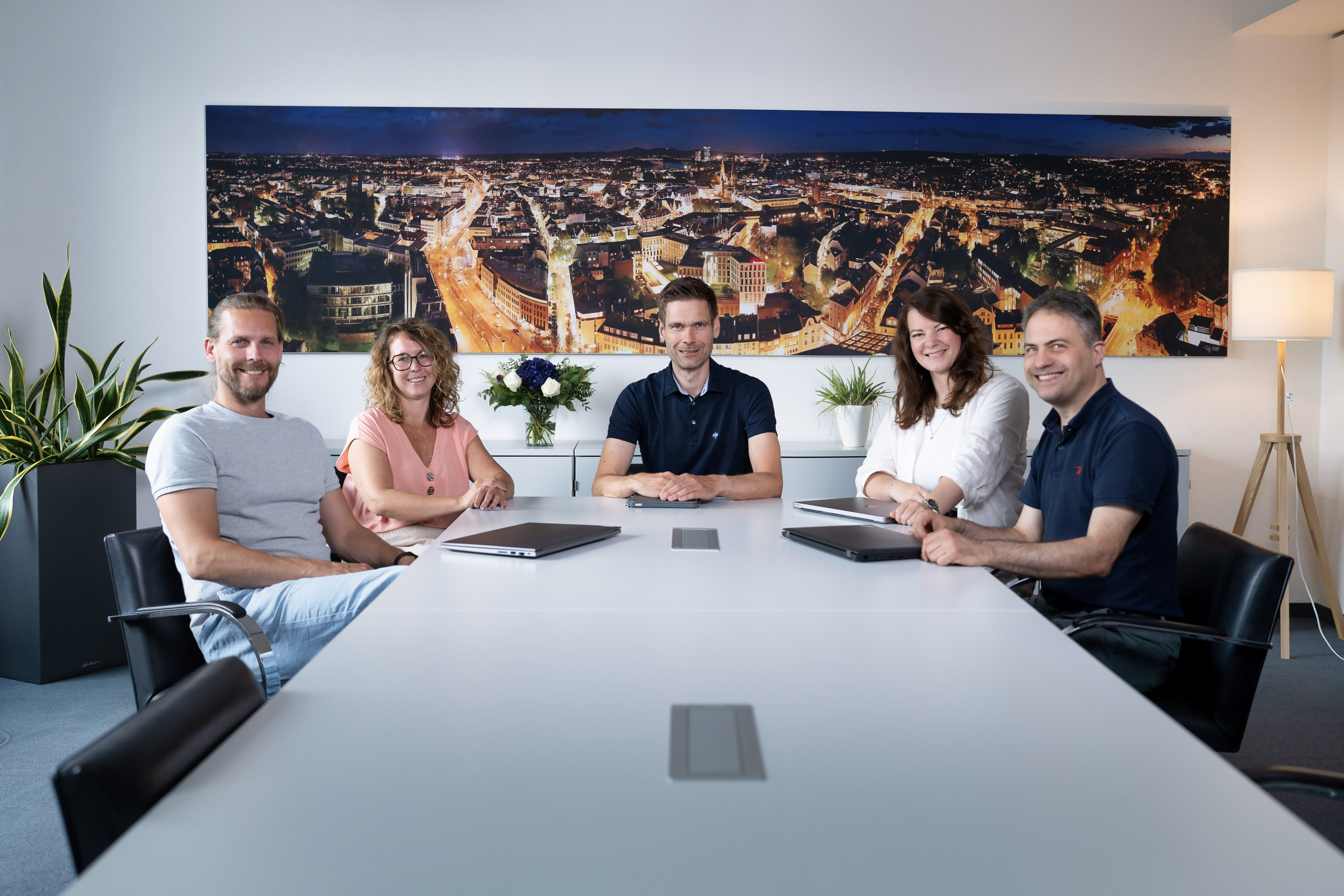 Fünf Personen sitzen an einem großen Tisch in einem modernen Konferenzraum, umgeben von Pflanzen und mit einer Stadtansicht im Hintergrund. Sie schauen lächelnd in die Kamera.