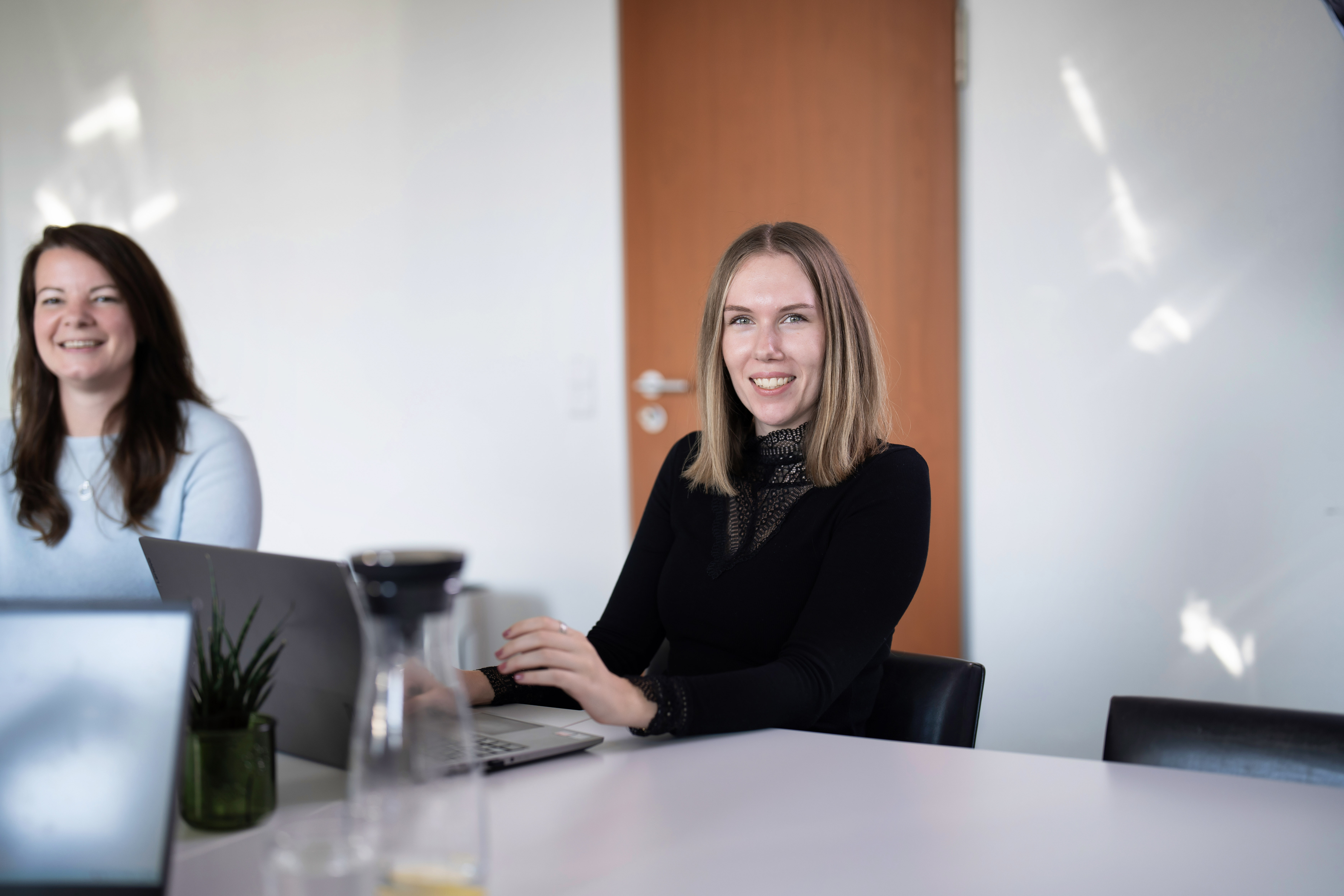 Zwei Frauen sitzen an einem Tisch in einem hellen Büroraum, während die Frau rechts lächelnd in die Kamera schaut und an einem Laptop arbeitet. Auf dem Tisch steht eine Pflanzen dekorative und ein Glaskaraffe.