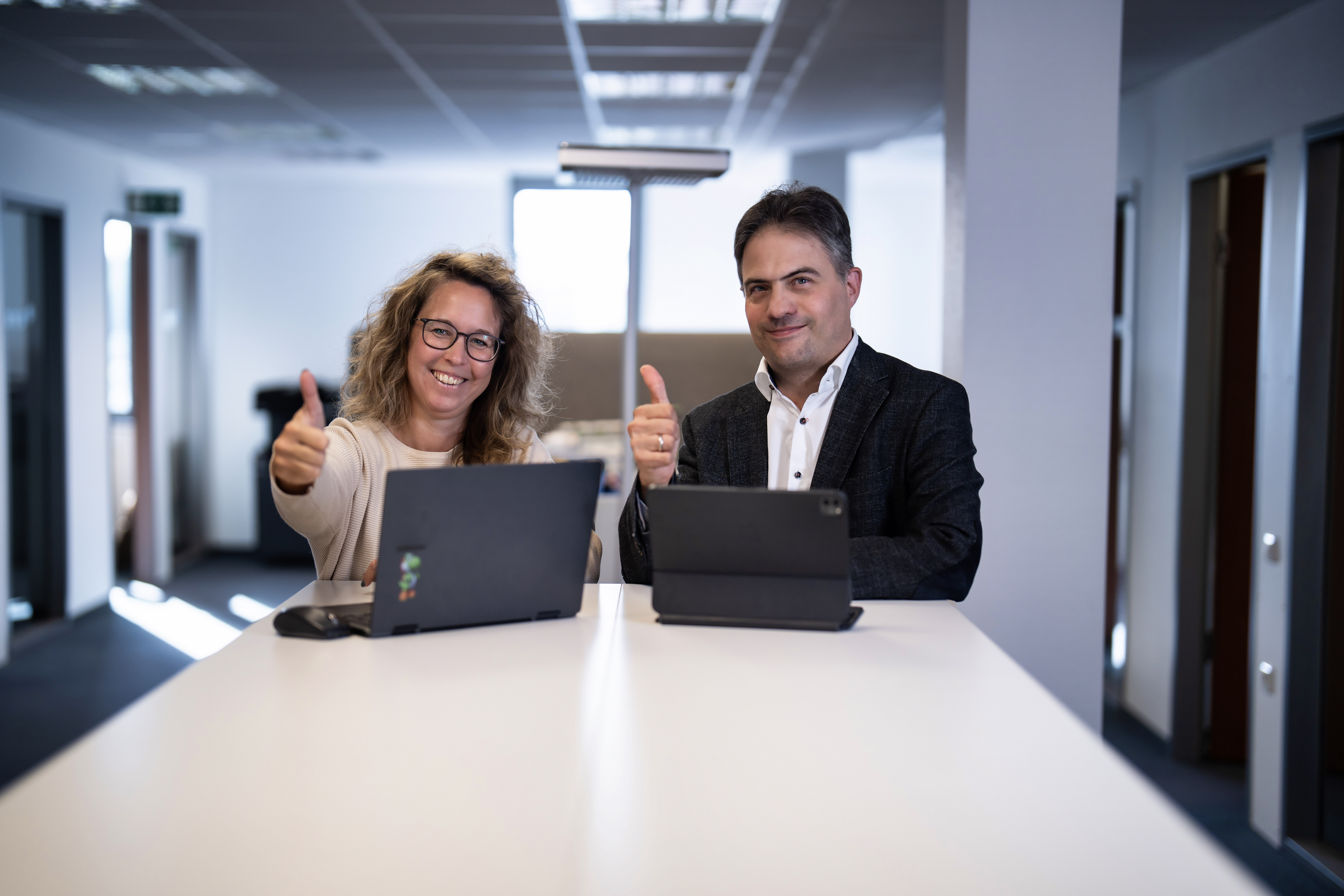 Eine Frau und ein Mann sitzen an einem Tisch in einem Büro, beide zeigen mit Daumen nach oben, während sie auf ihre Laptops schauen.