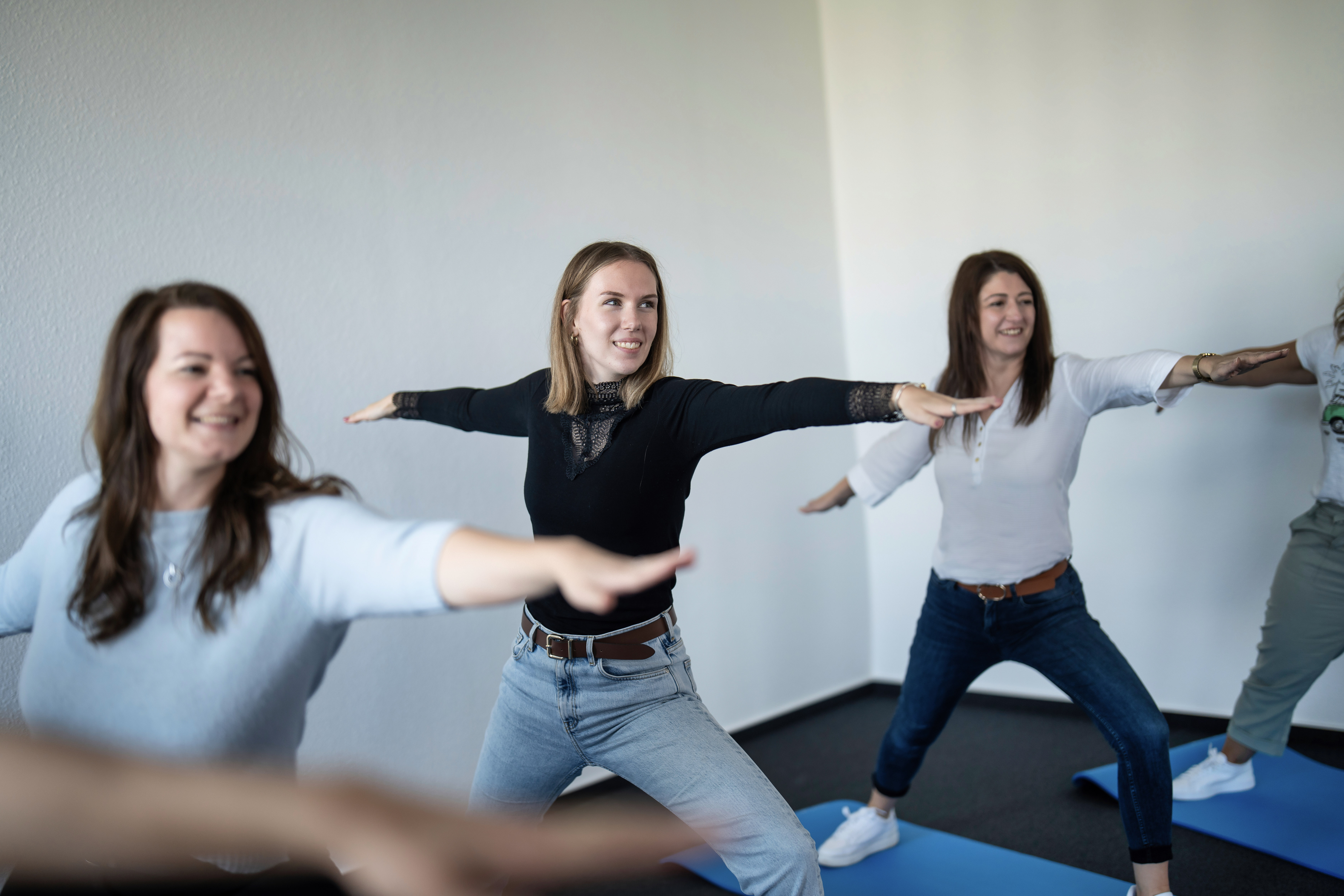 Drei Frauen führen gemeinsam eine Yoga-Übung in einer hellen, modernen Umgebung durch, wobei sie in einer Krieger-Pose stehen und ihre Arme zur Seite ausstrecken.