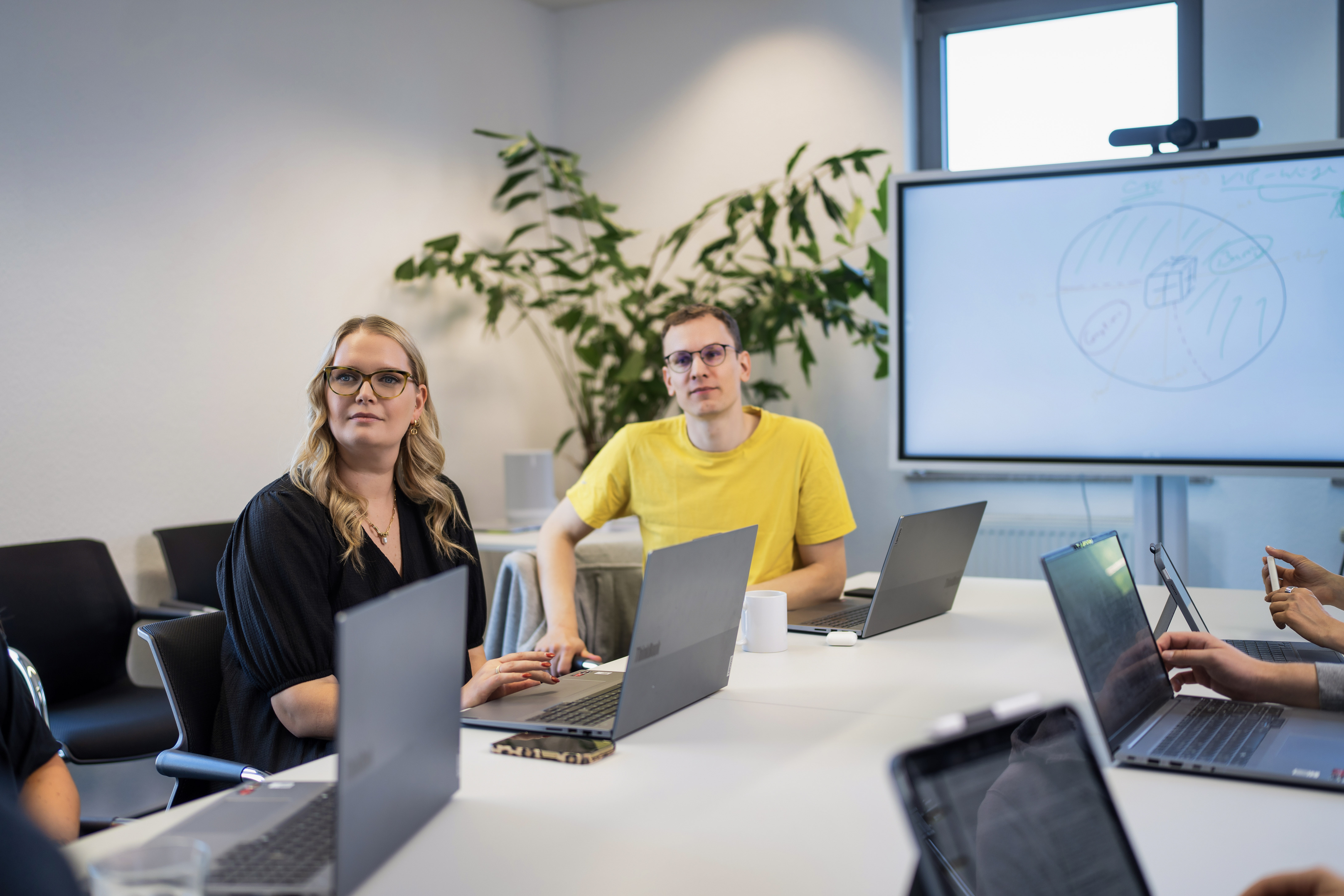 Eine Gruppe von Personen sitzt um einen Tisch in einem Konferenzraum, während sie an Laptops arbeiten und einem Bildschirm mit einer Präsentation im Hintergrund folgen. Zwei der Personen, eine Frau mit langen Haaren und Brille sowie ein Mann mit kurzen Haaren in einem gelben T-Shirt, schauen aufmerksam in die Richtung des Sprechers. In der Ecke des Raumes steht eine große Pflanze.