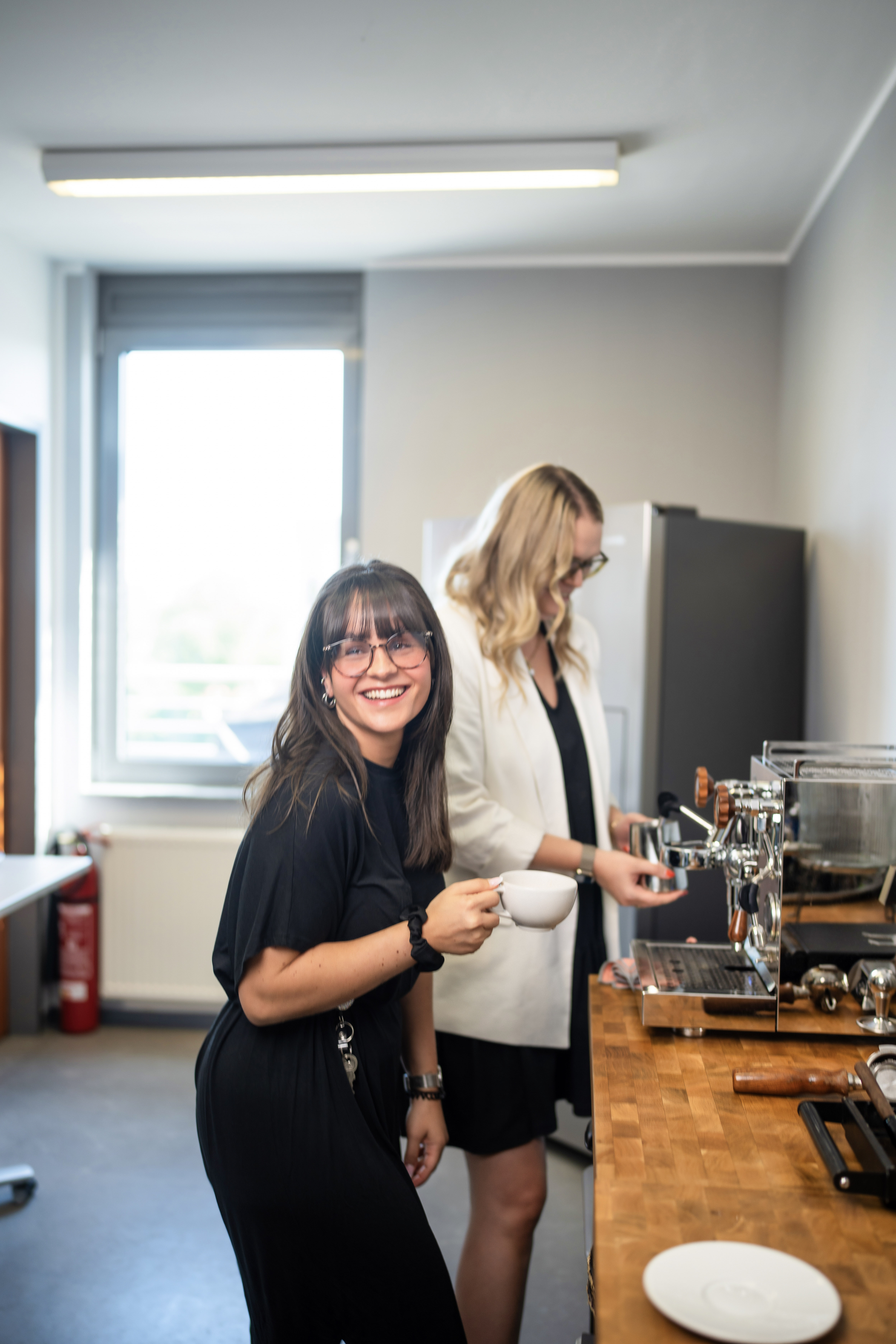 Eine Frau in schwarzer Kleidung lächelt und hält eine Kaffeetasse in der Hand, während sie sich in einer modernen Küche befindet. Im Hintergrund steht eine weitere Frau in einem weißen Blazer, die an einer Espressomaschine arbeitet. Tageslicht strömt durch das Fenster.
