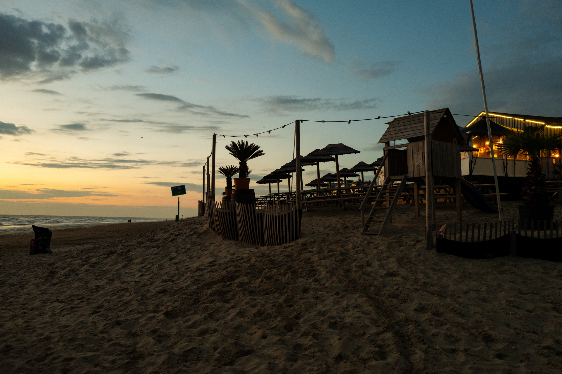 Eine Strandlandschaft bei Sonnenuntergang, mit einer Spielstruktur aus Holz, Liegestühlen unter Sonnenschirmen und einem gemütlichen Restaurant im Hintergrund, umgeben von sandigem Gelände und spärlicher Beleuchtung.
