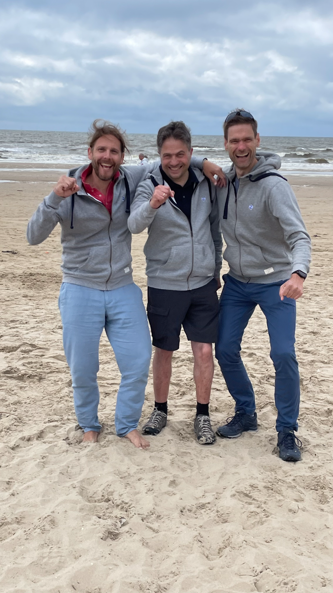 Drei Männer in grauen Kapuzenpullovern stehen am Strand, lächeln und zeigen Daumen hoch, während sie vor dem Meer pic.twitter.com. Der Himmel ist bewölkt und der Sandstrand ist gut sichtbar.