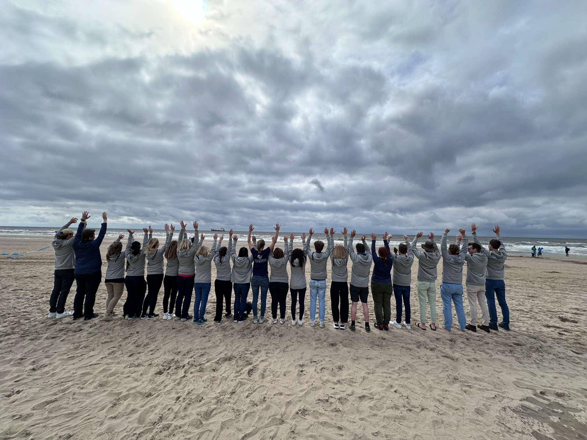 Eine Gruppe von Menschen steht am Strand mit den Händen in die Höhe, während sie in Richtung des Meeres blicken. Der Himmel ist bewölkt und es gibt einen leicht grauen Sandstrand.