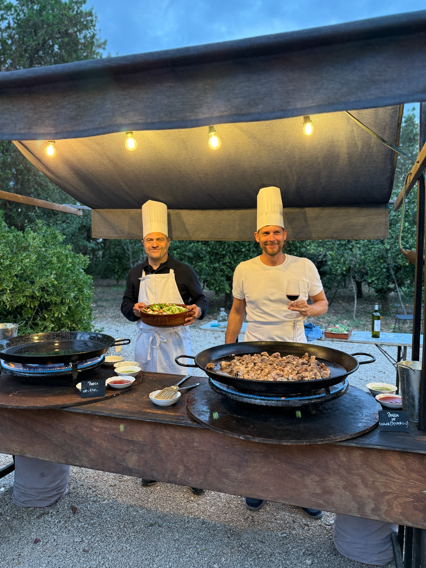 Zwei Köche in Kochmützen stehen an einem Freiluftküchenstand, der mit drei großen Pfannen mit Essen ausgestattet ist. Einer der Köche hält eine Schüssel mit Salat, während der andere ein Glas Wein in der Hand hält. Über ihnen hängen Lichter und im Hintergrund sind Bäume zu sehen.