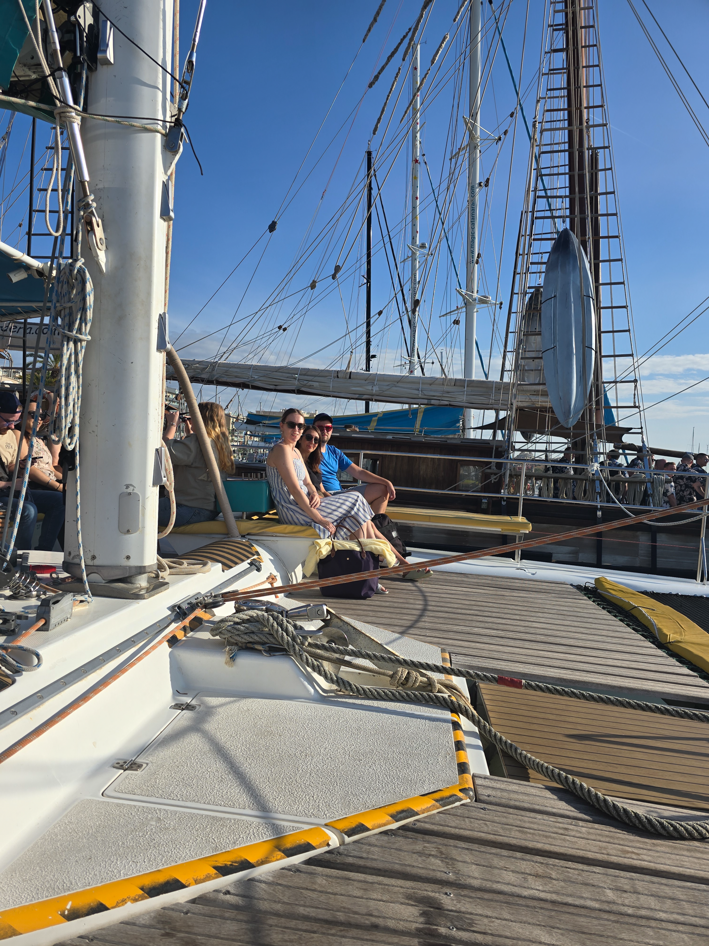 Zwei Personen sitzen an Bord eines Segelbootes im Hafen, umgeben von anderen Booten und einem klaren blauen Himmel.