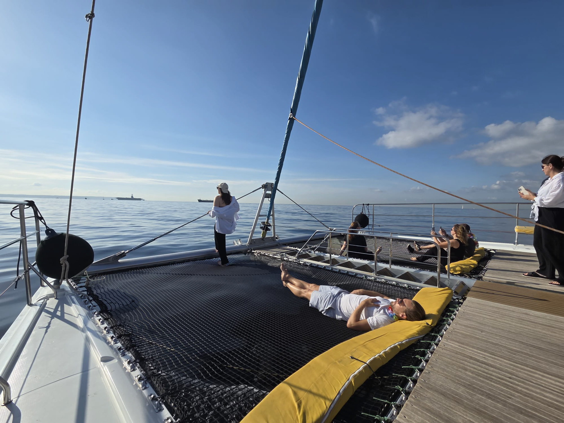 Personen entspannen auf einem Katamaran, während sie die ruhige Meereslandschaft und den blauen Himmel genießen. Eine Person liegt auf einem großen Netz vorne am Boot, während andere stehen und sitzen, um die Aussicht zu genießen.