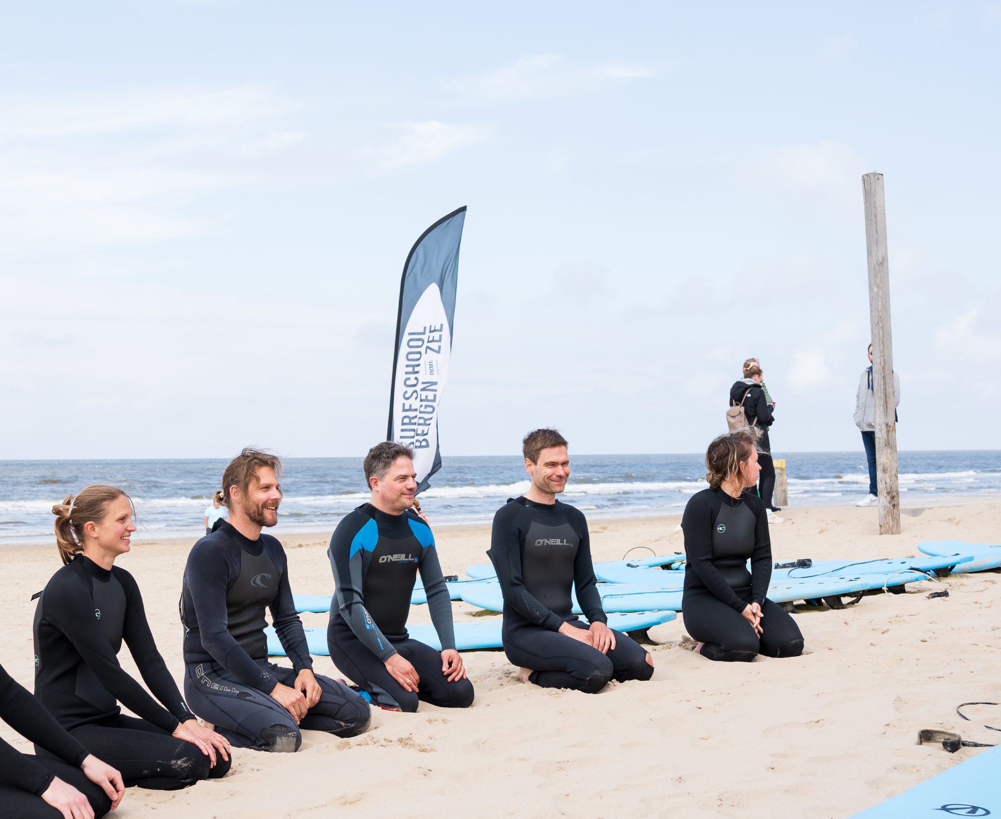 Eine Gruppe von Personen in Neoprenanzügen sitzt am Strand und hört einem Surf-Lehrer zu, während im Hintergrund das Meer und ein Surfbrett-Flaggenbanner zu sehen sind.