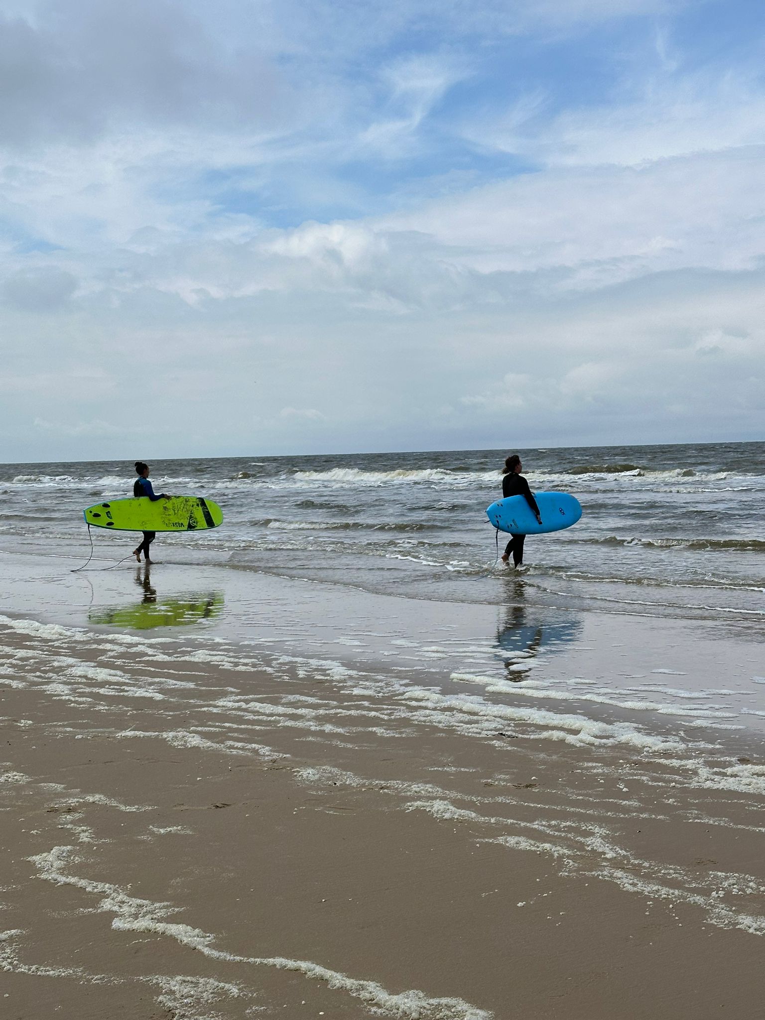 Zwei Surfer gehen mit ihren Surfbrettern, einem grünen und einem blauen, am Strand entlang ins Wasser, während die Wellen sanft an das Ufer rollen und der Himmel bewölkt ist.